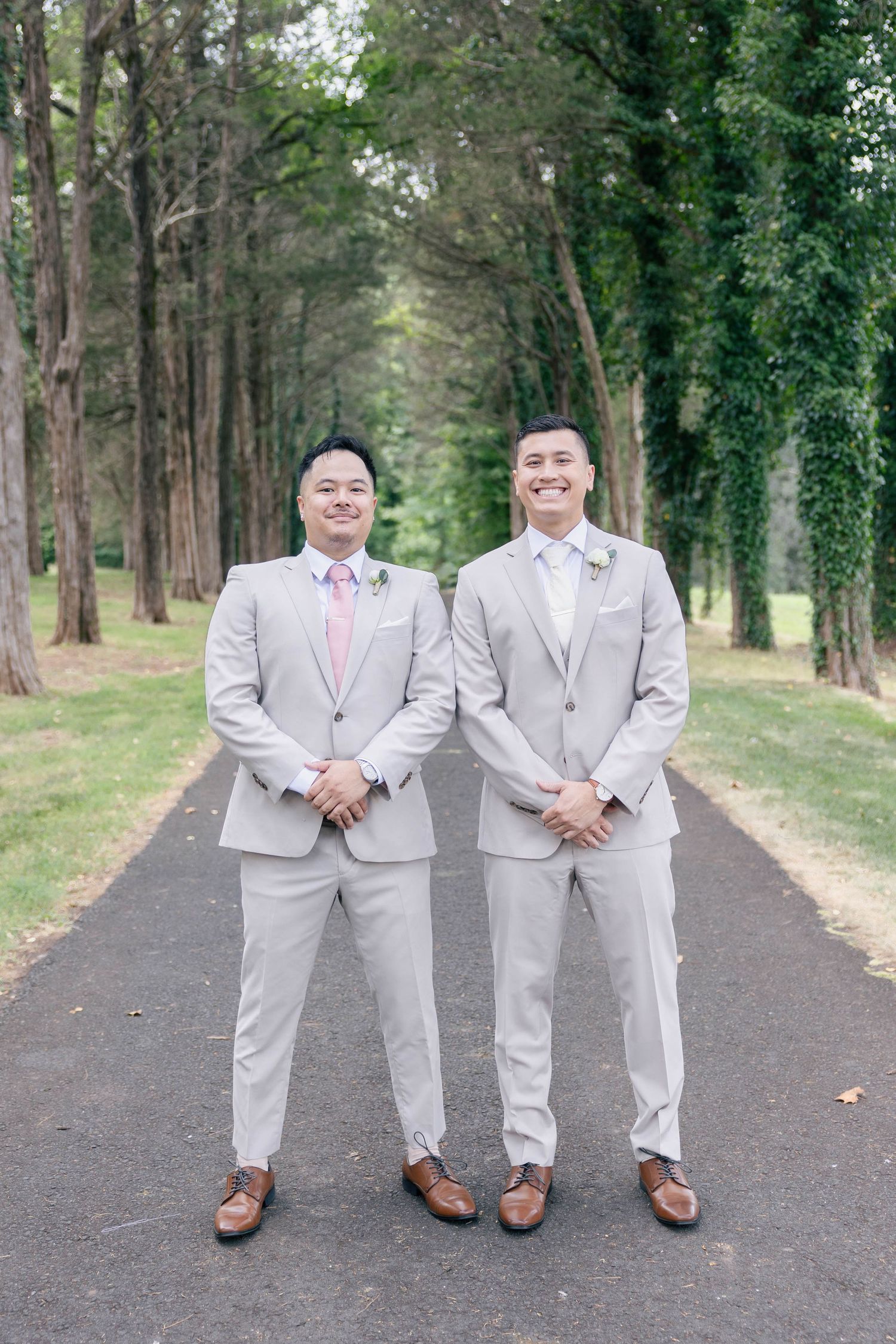 Wedding party members in light gray suits pose together on wooded pathway. Groom and groomsmen wedding outfit inspiration in tan suit.