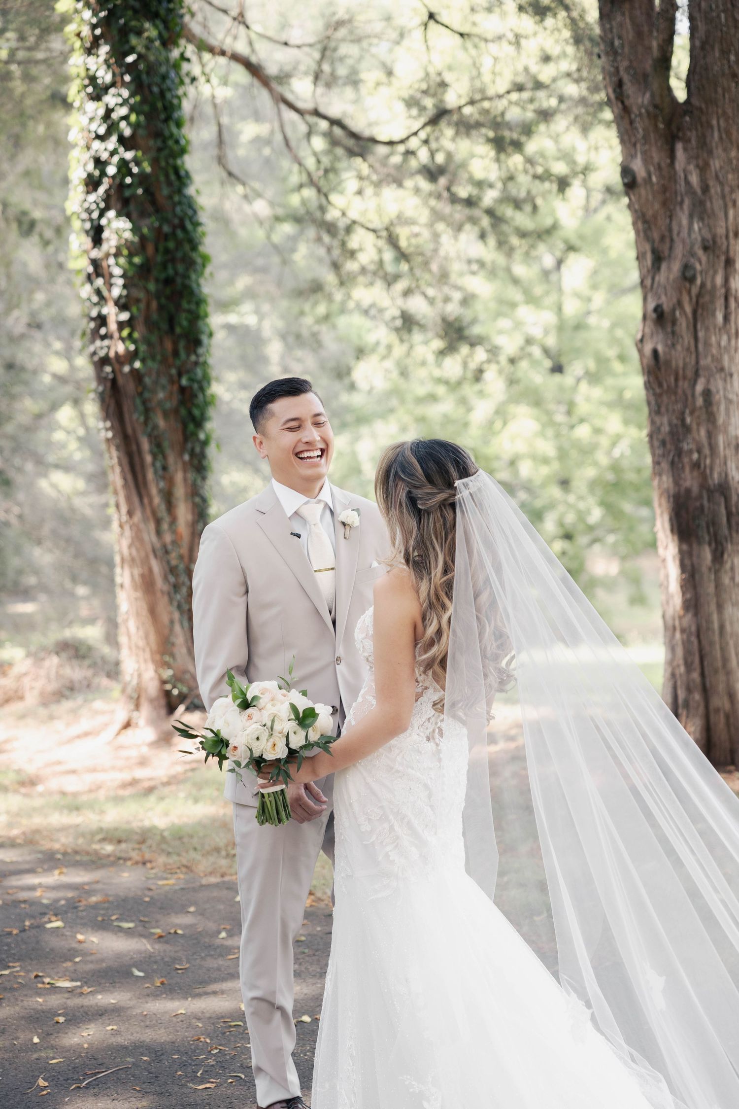 A wedding couple laughs together during their first look in a wooded setting.