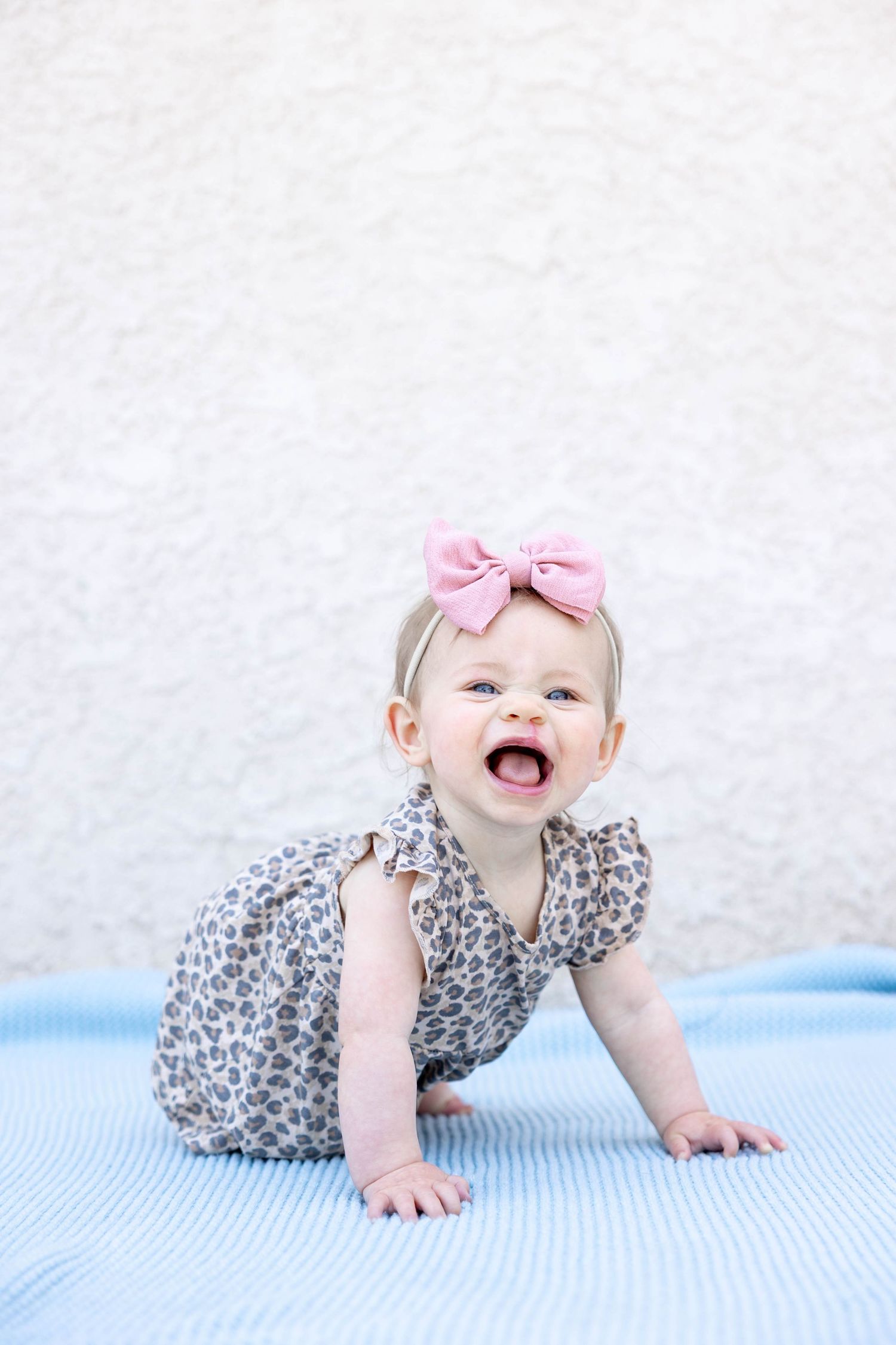 Baby wearing leopard print dress and pink bow crawls on blue mat while laughing joyfully.