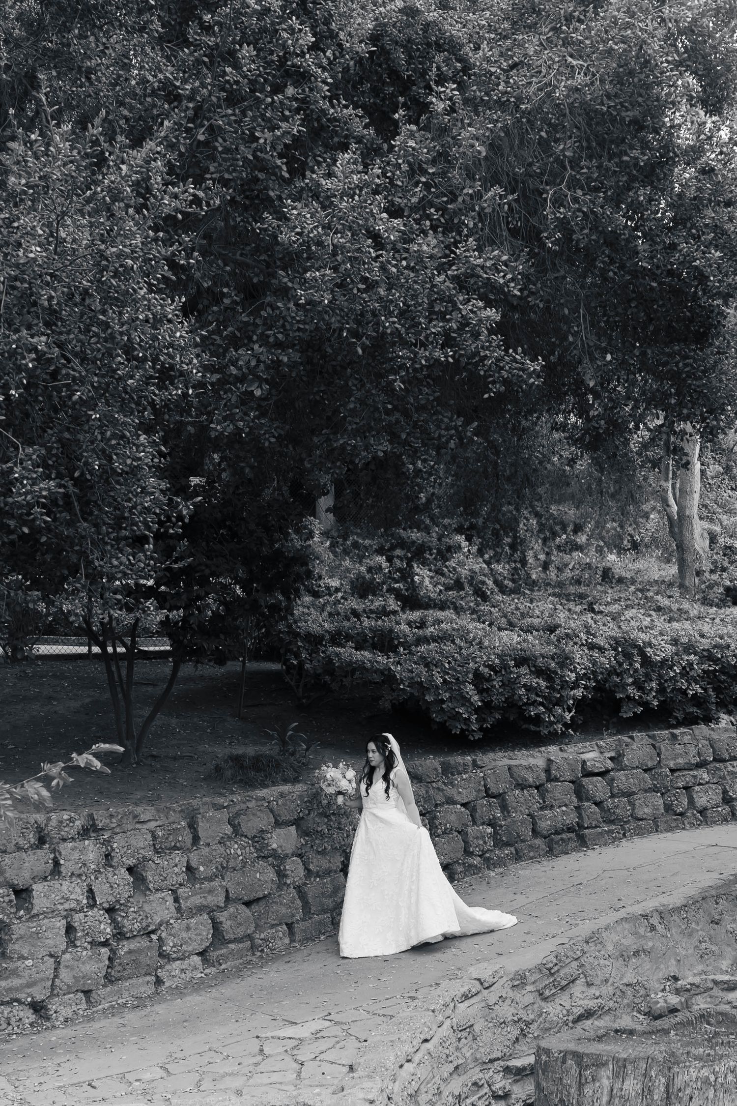 A black and white wedding photo series showing a couple sitting on a stone wall in a garden setting.