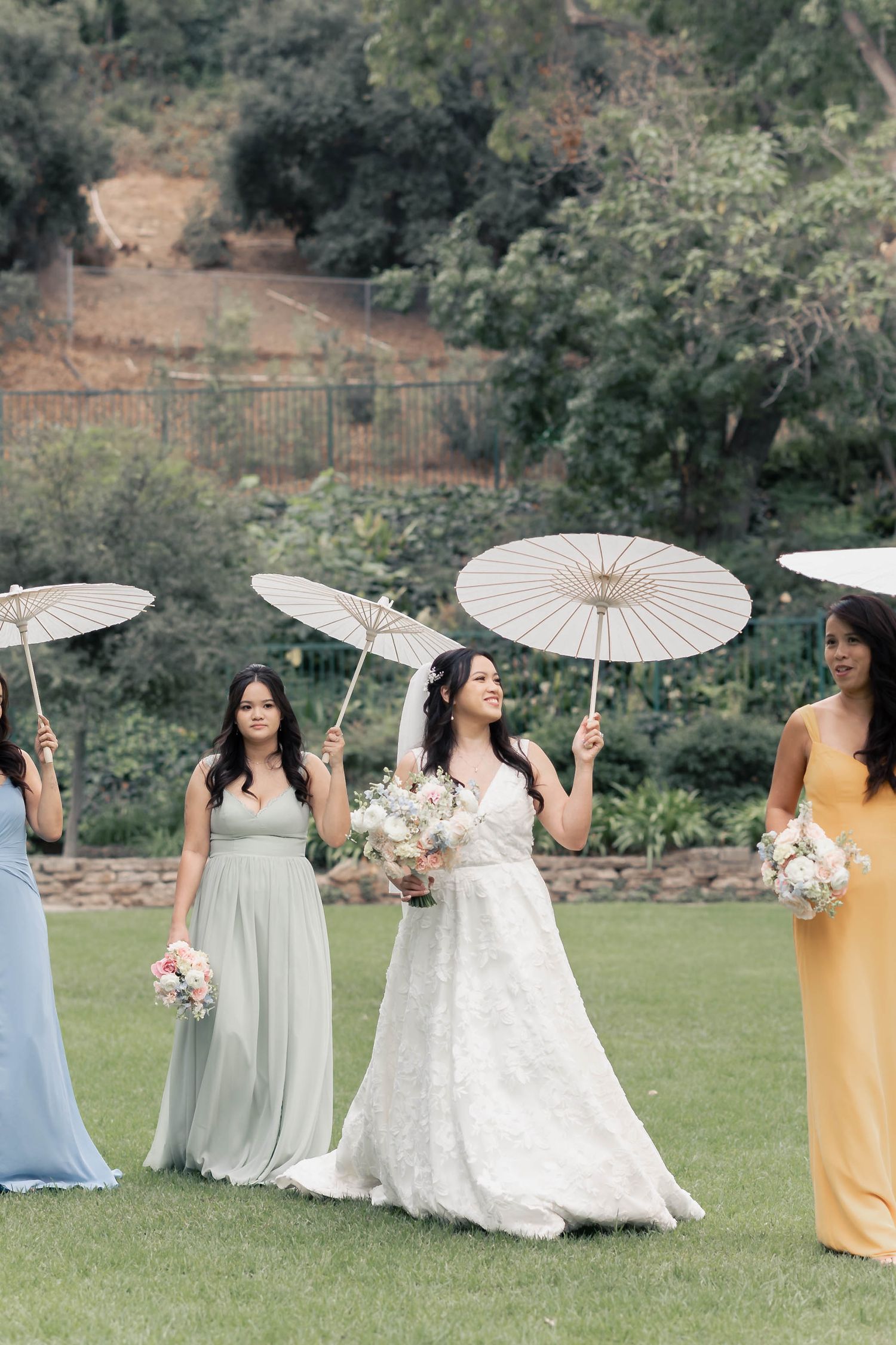 A wedding party poses with elegant white parasols on a grassy lawn with colorful bridesmaid dresses.