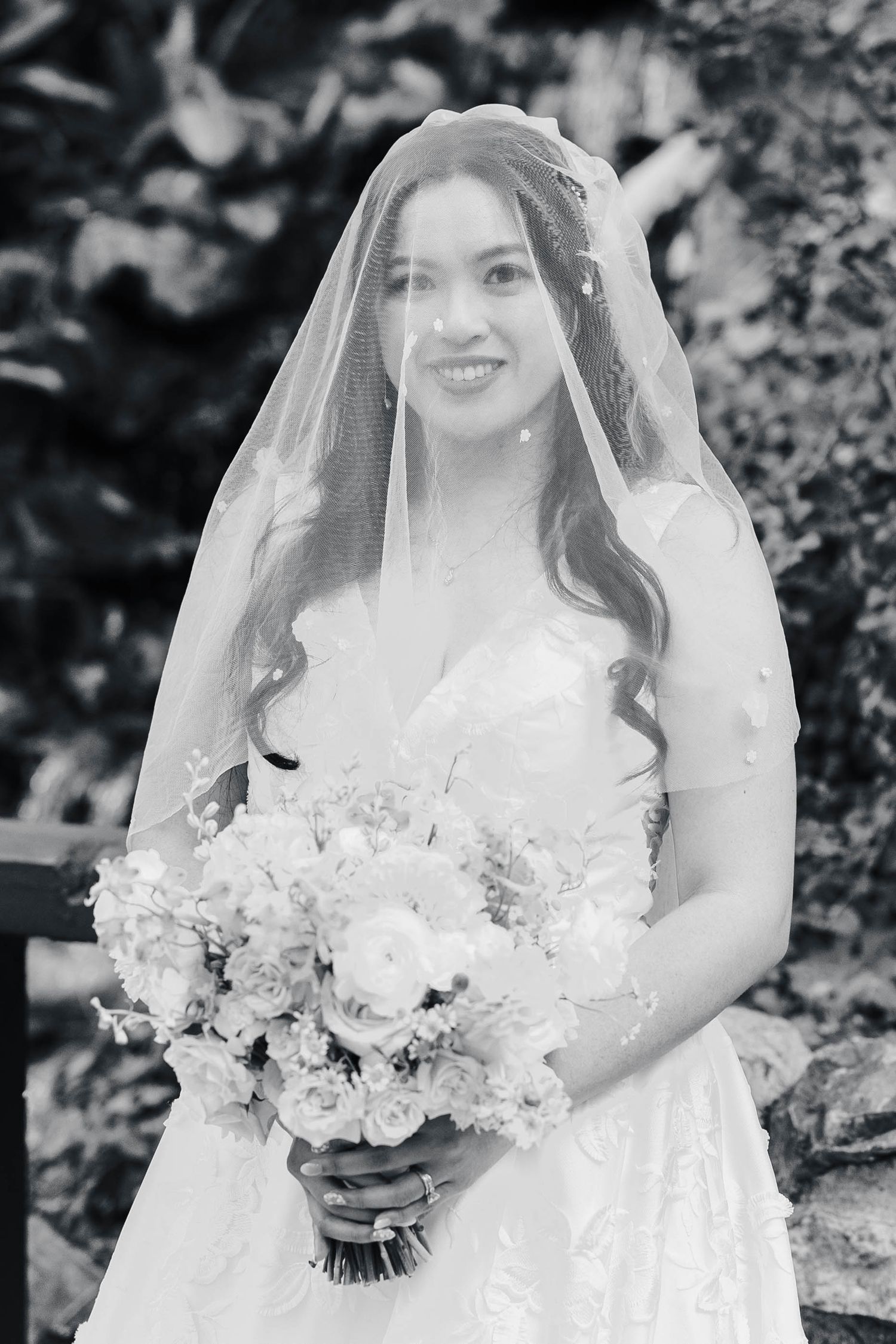 Black and white portrait of someone in a wedding dress and veil holding a bouquet of white flowers.