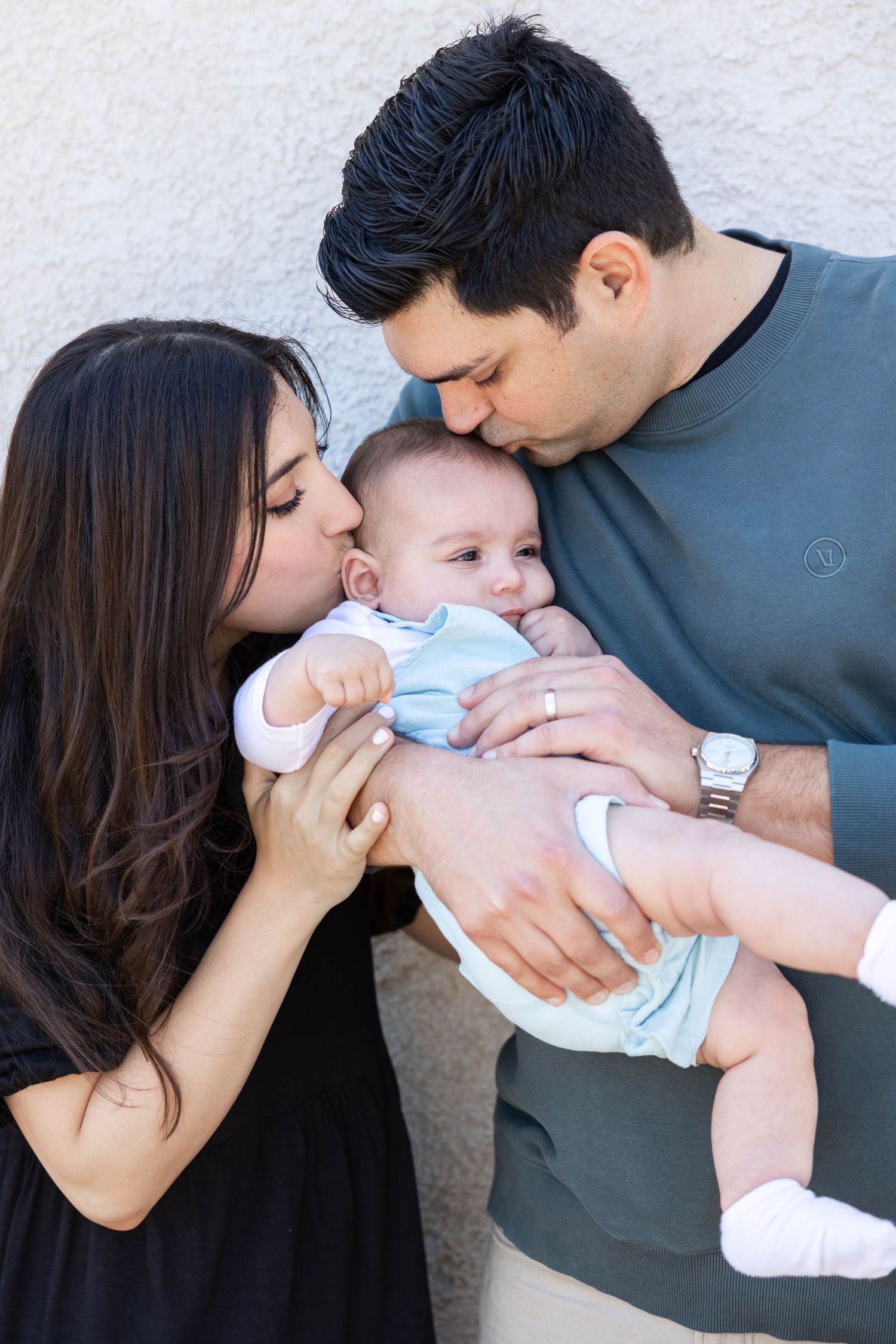 Three connected images show tender family moments as parents embrace and kiss their baby wearing a light blue outfit.