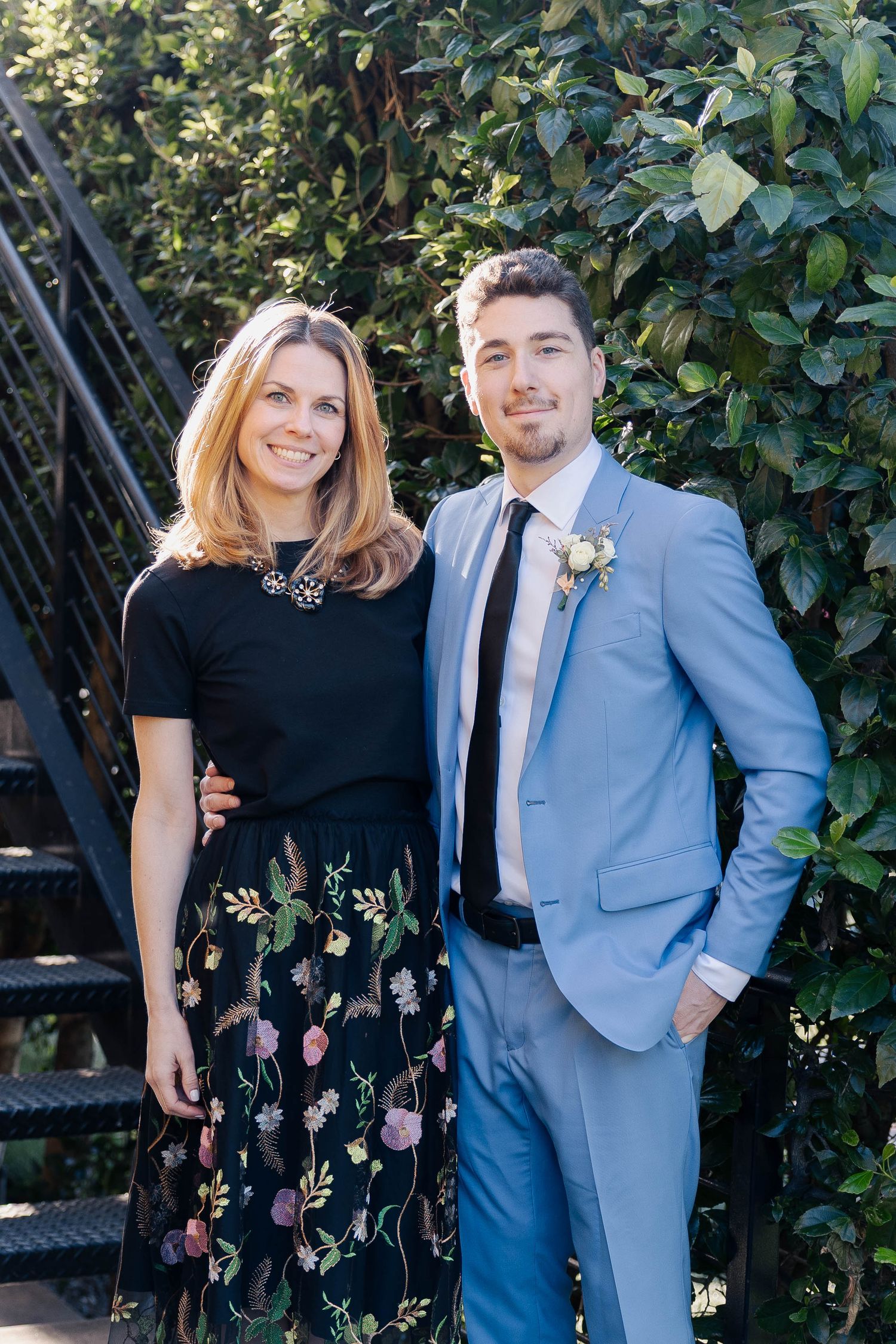 Outdoor portrait of two people against iron staircase and green foliage.