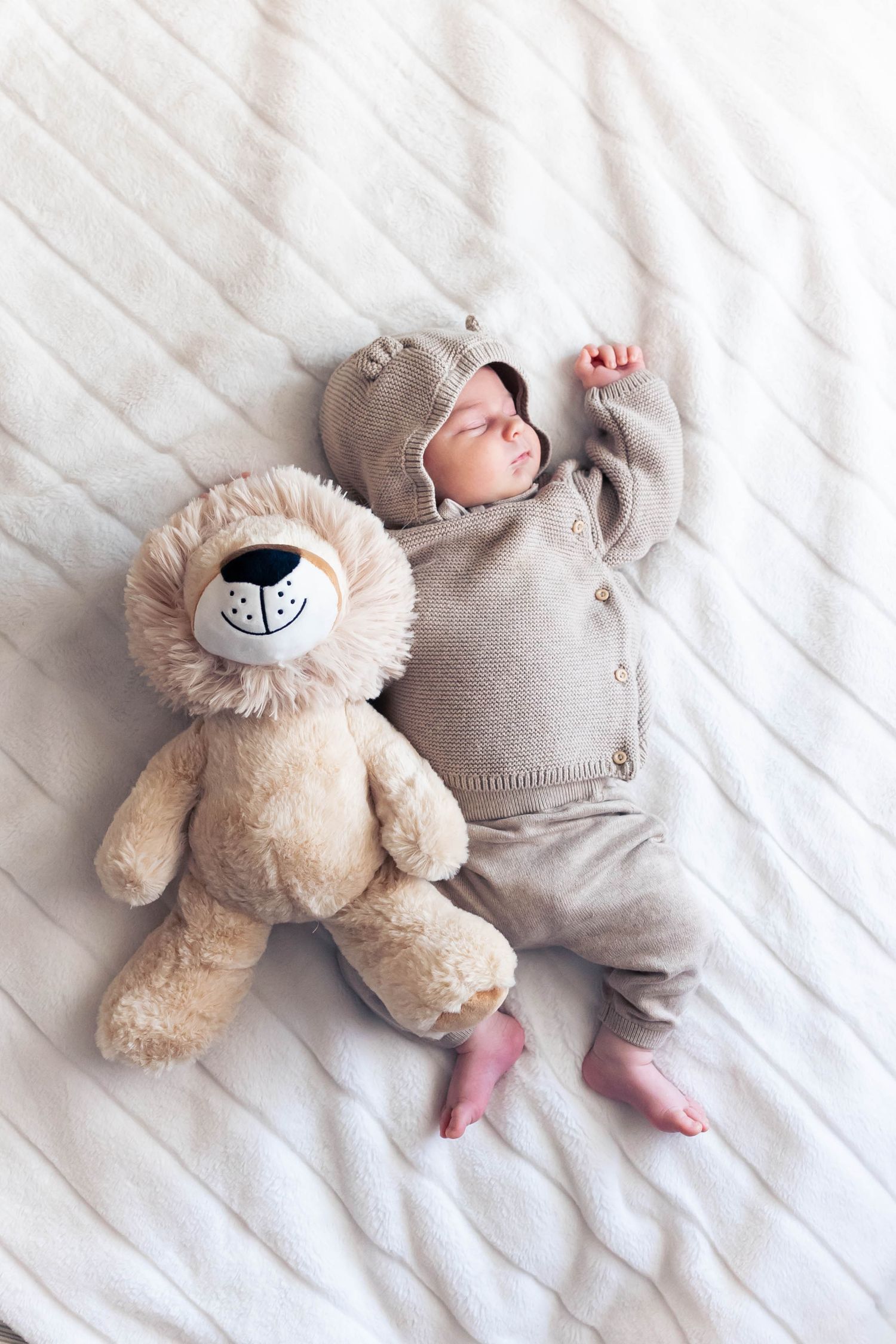 A small infant lies peacefully on white bedding next to a soft plush lion toy.