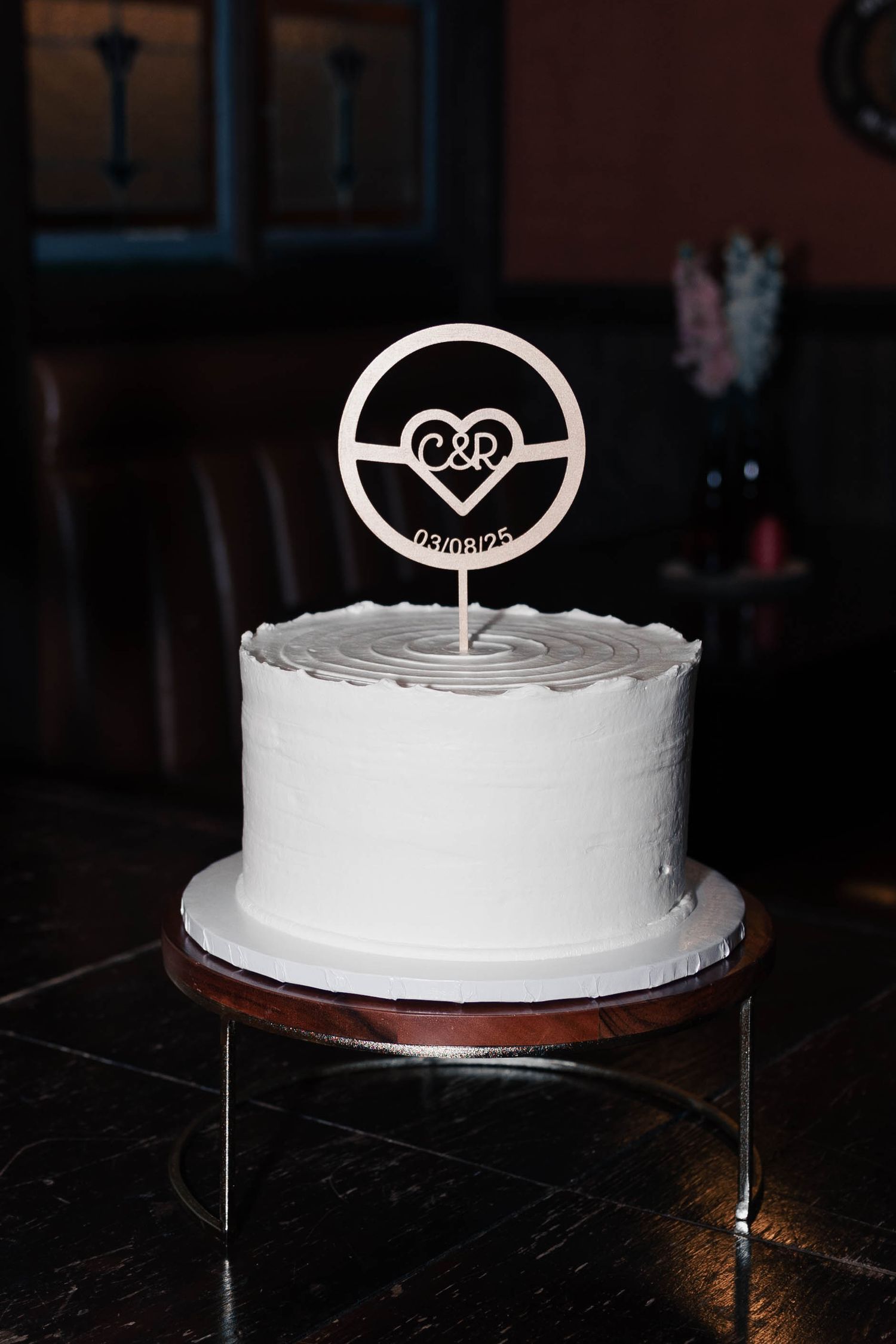 A white frosted wedding cake with a heart-shaped topper displayed on a dark surface from multiple angles.