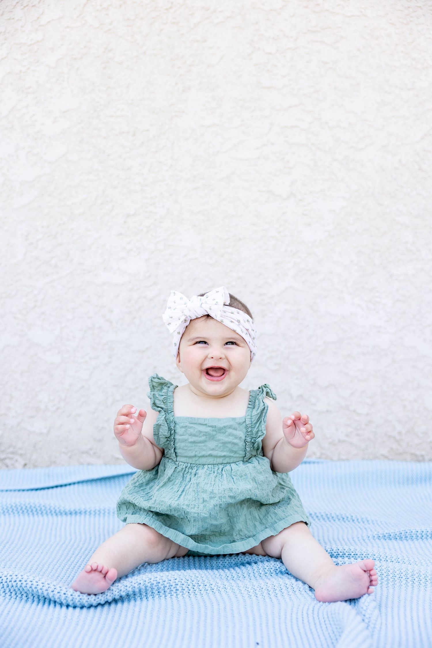 Happy baby in mint green dress sitting on striped blanket against white wall.