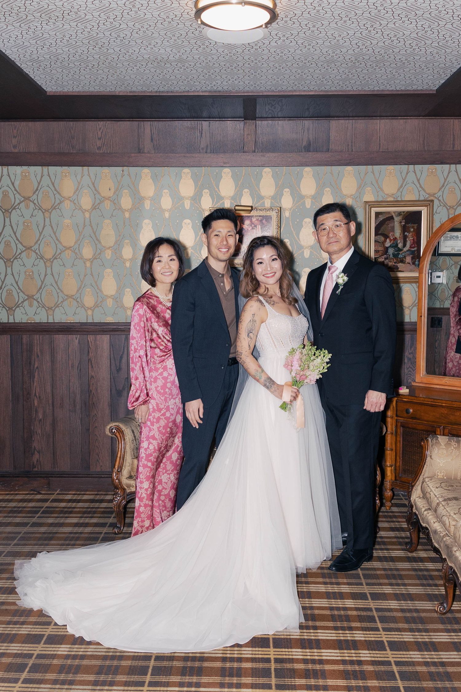 Wedding group photo in formal attire against wood-paneled walls and vintage wallpaper.