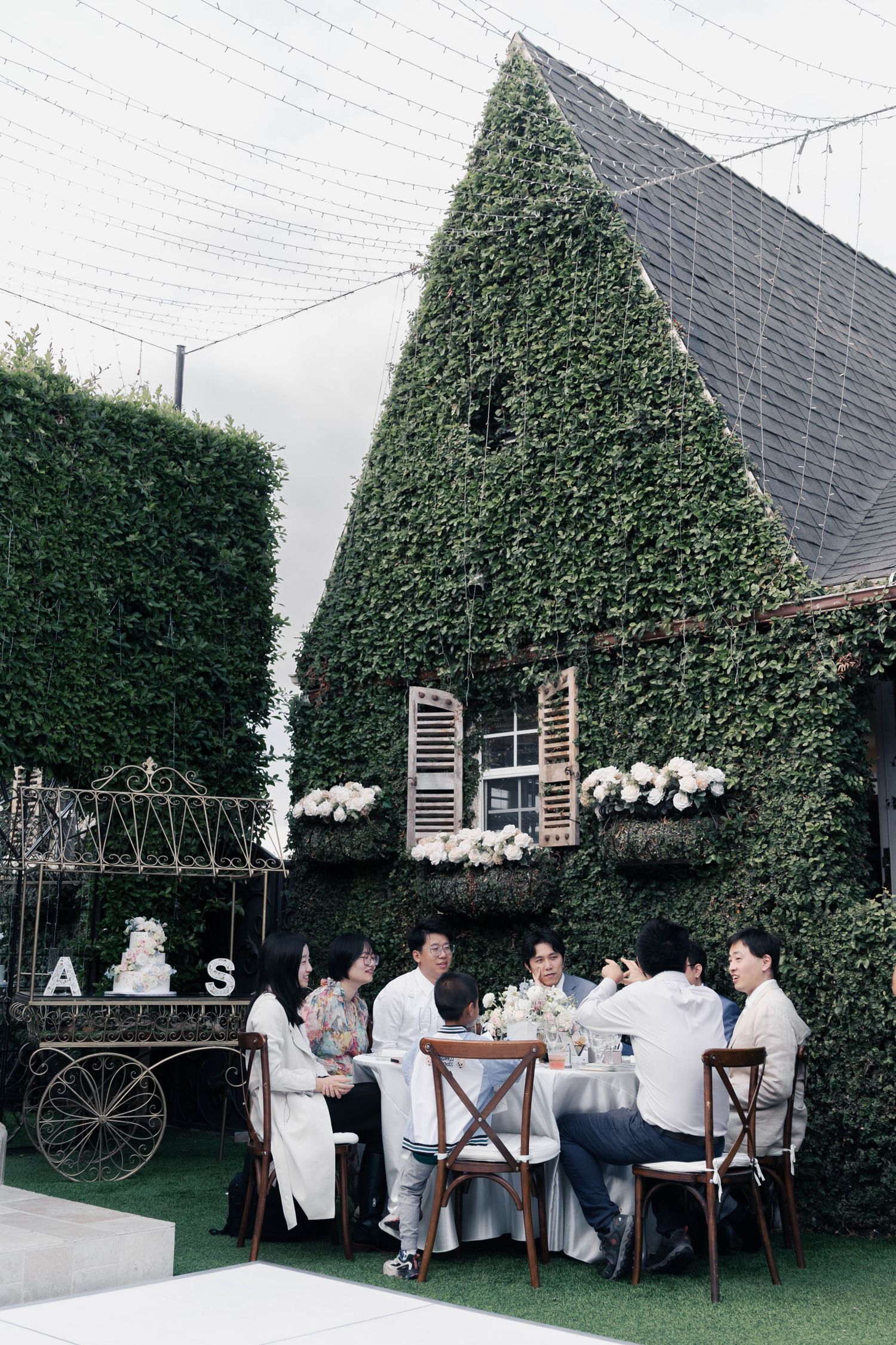 Outdoor wedding reception setup with long table against green hedge wall and elegant white decor details.