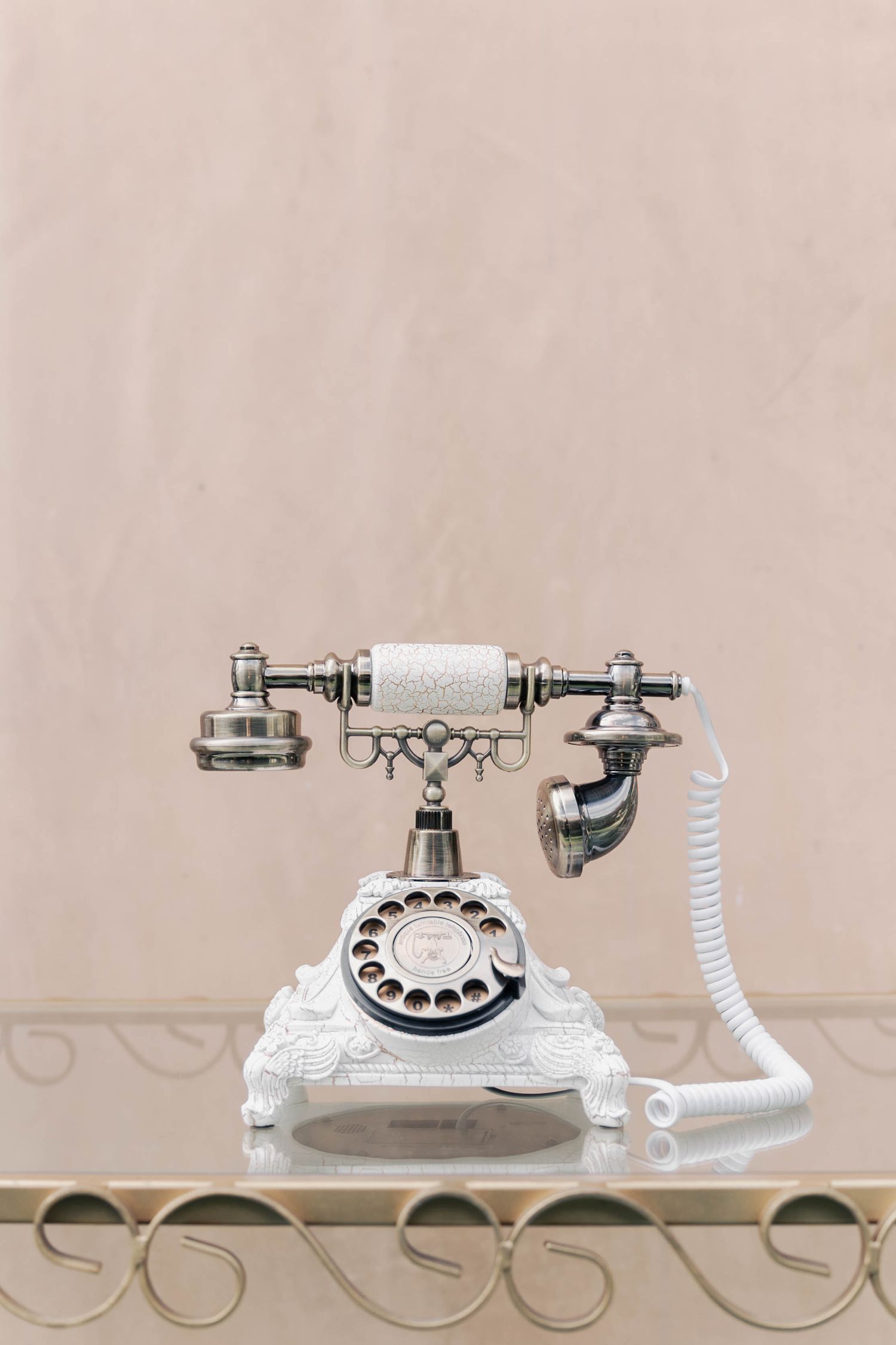 Vintage white ornate rotary telephone with decorative metallic details sits elegantly on a curved metal table.