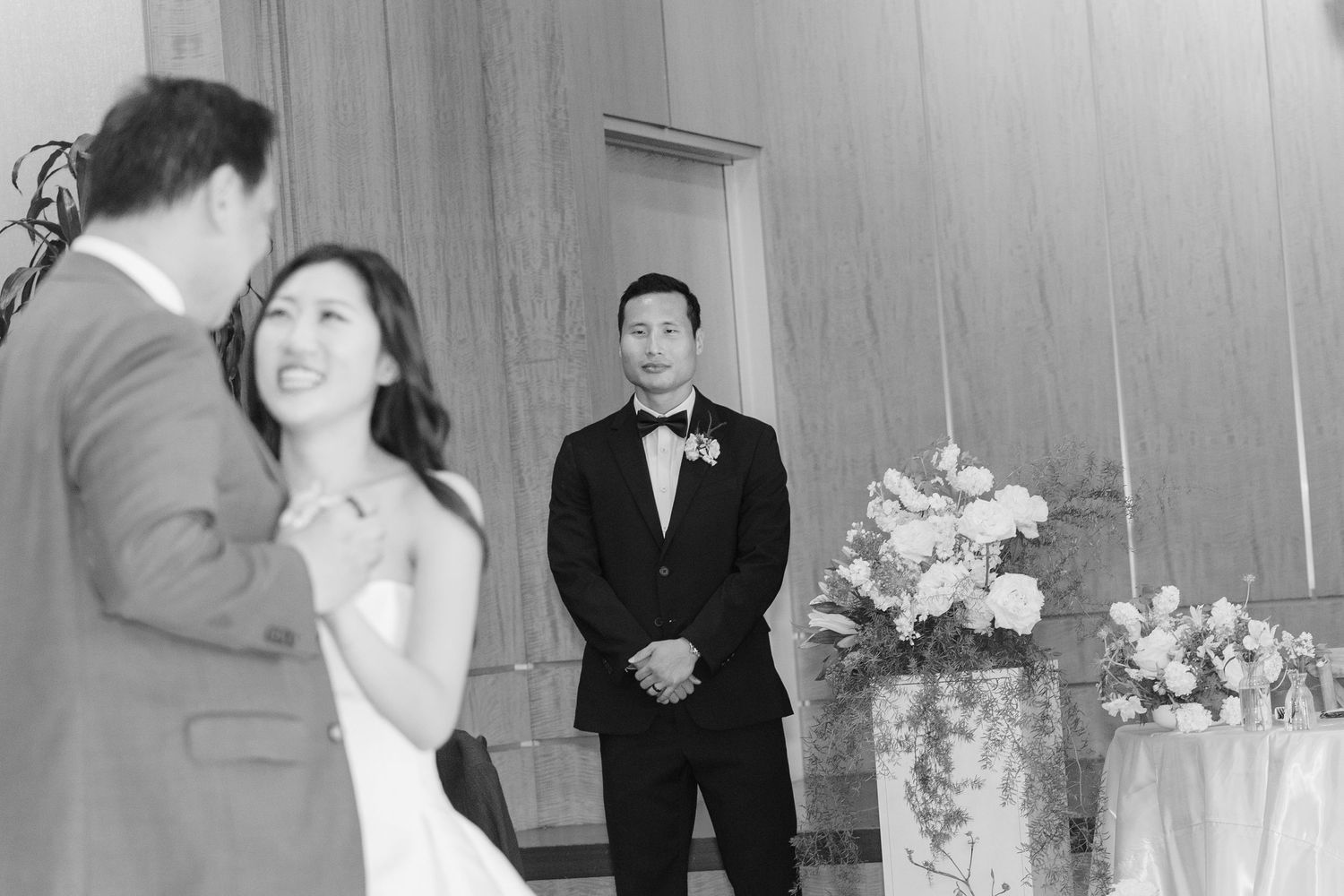Black and white wedding photo of bride and groom sharing a moment as a guest looks on near floral arrangements.