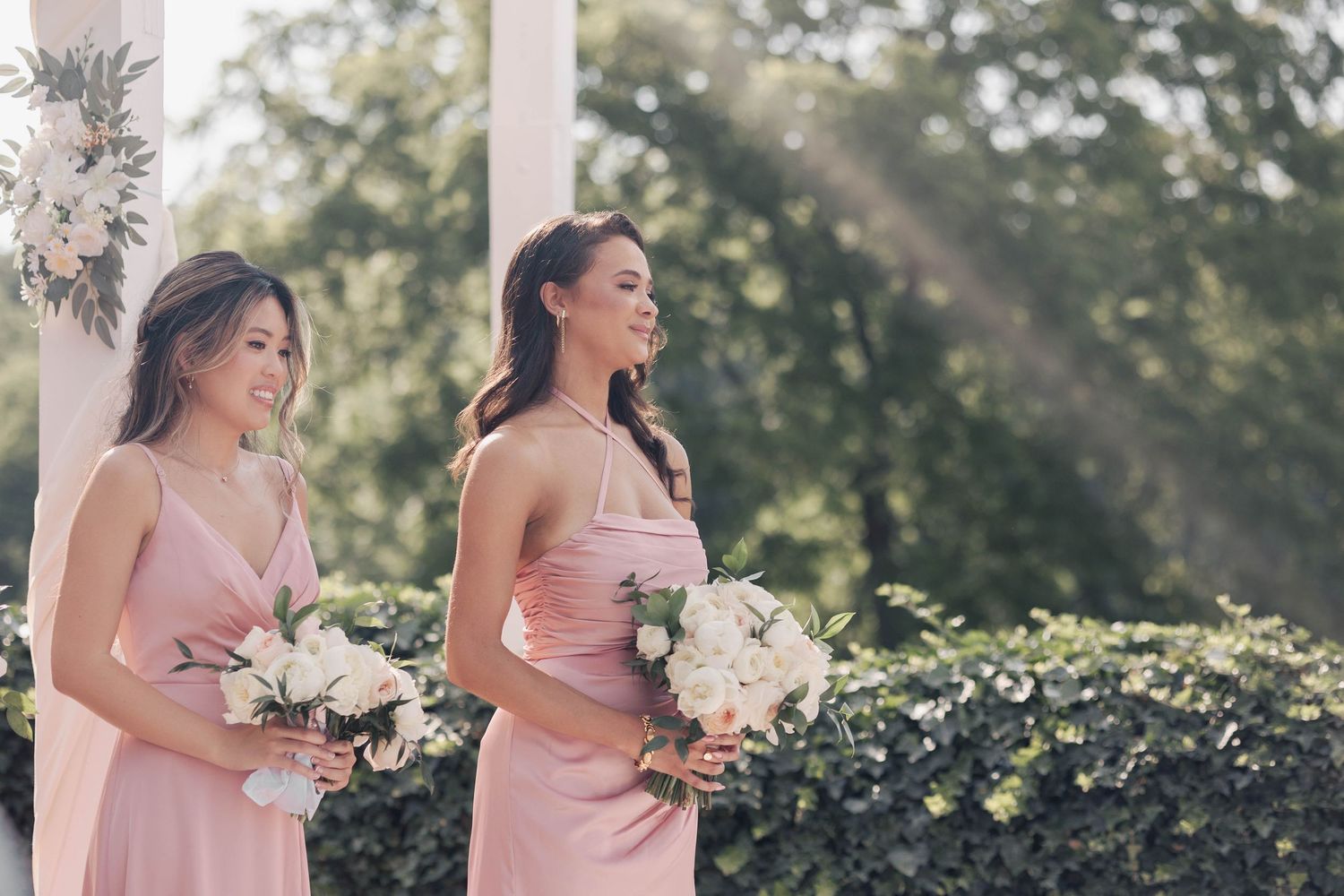 Bridesmaids in blush pink dresses hold white bouquets while smiling outdoors at a wedding ceremony.