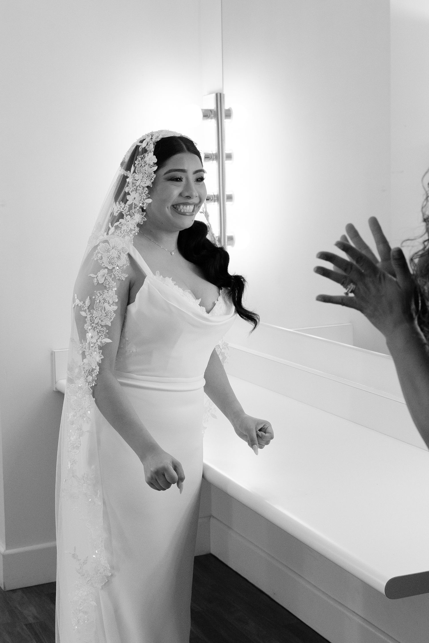 Smiling bride in white wedding gown and lace veil stands near bathroom vanity in black and white photo.