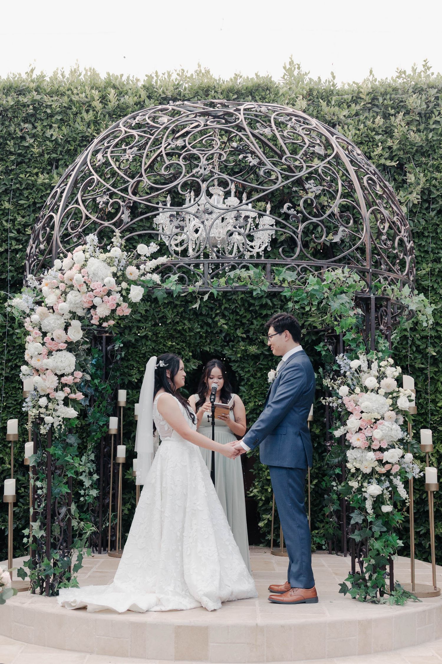 Elegant wedding ceremony under an ornate metal gazebo with crystal chandelier and blush pink floral arrangements.