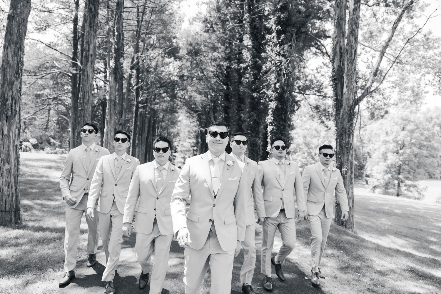A group in light gray suits walks along a tree-lined path during a wedding photoshoot.