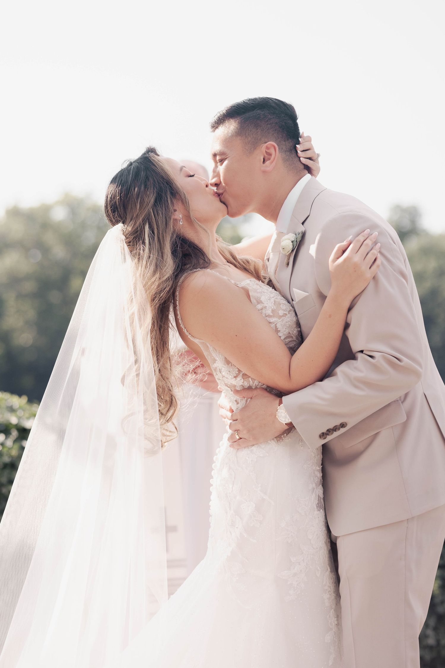 Newlyweds share an intimate first kiss under bright sunlight at their outdoor wedding ceremony.