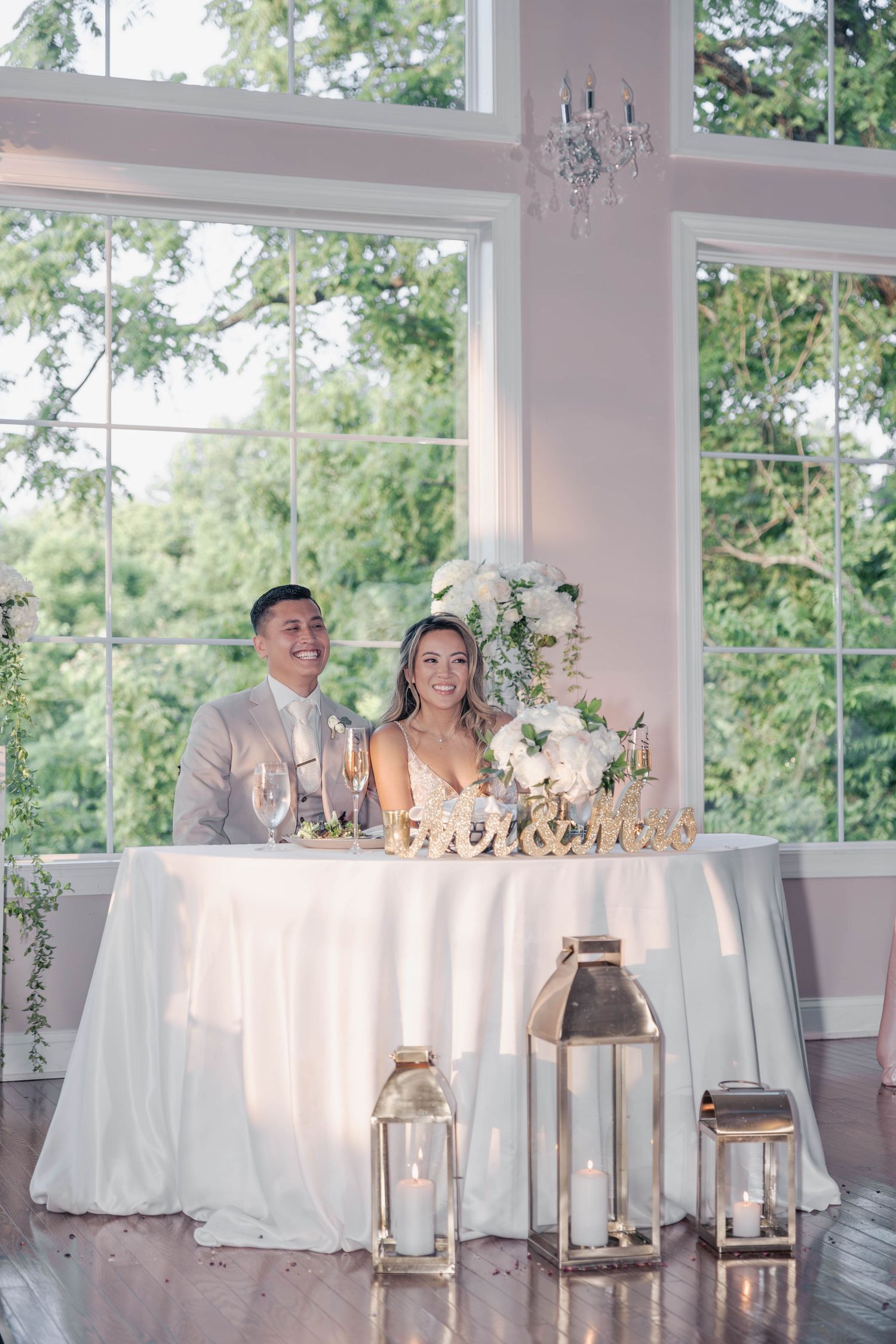 Wedding head table decorated with white linens and floral arrangements in a bright sunlit venue.