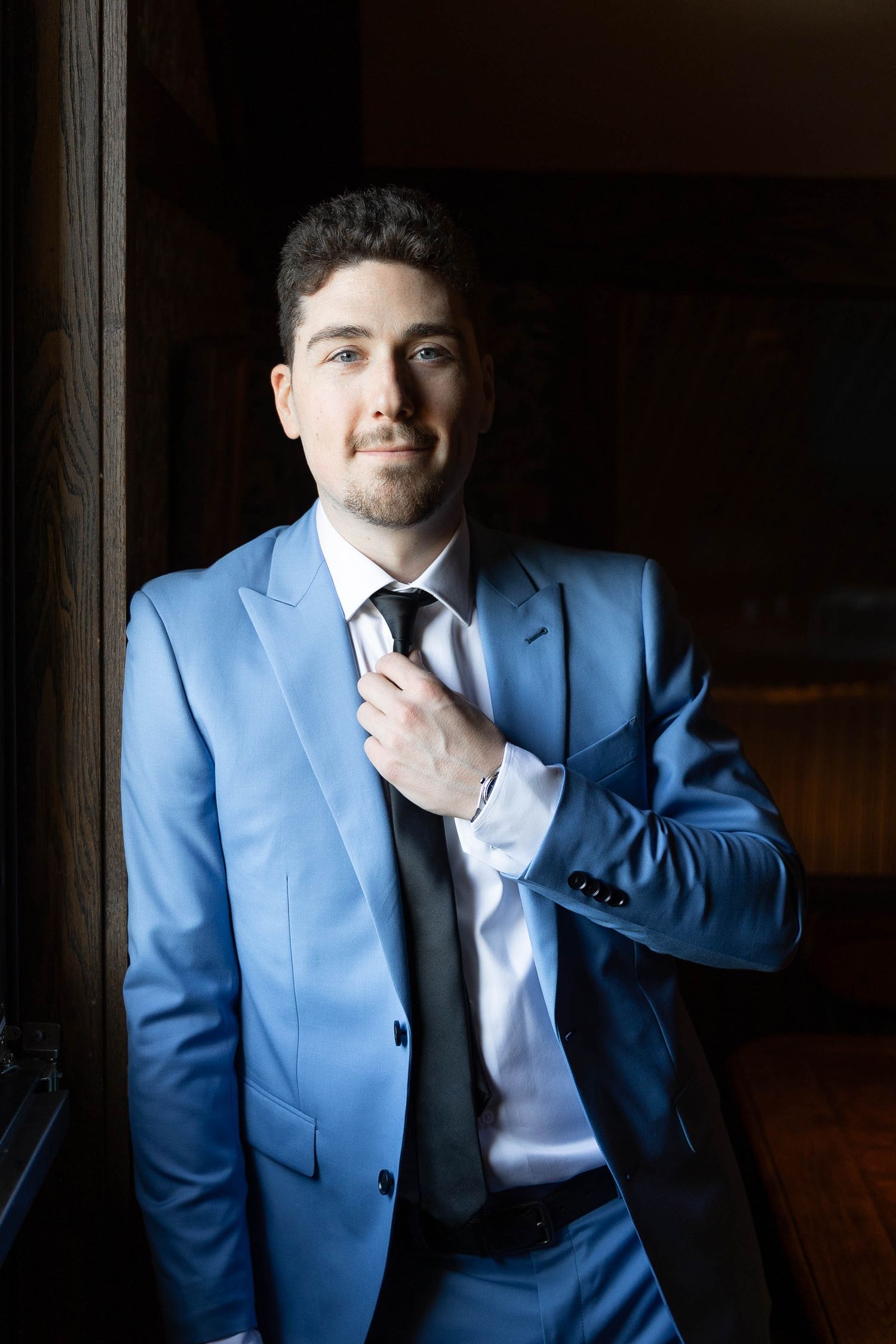 Person in a blue suit and tie stands in contemplative pose by a window in moody lighting.