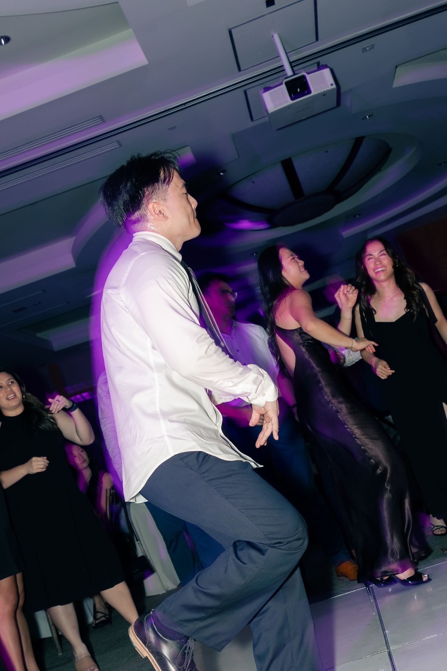 Man in white shirt dancing energetically at a party with guests in formal attire under purple lights.