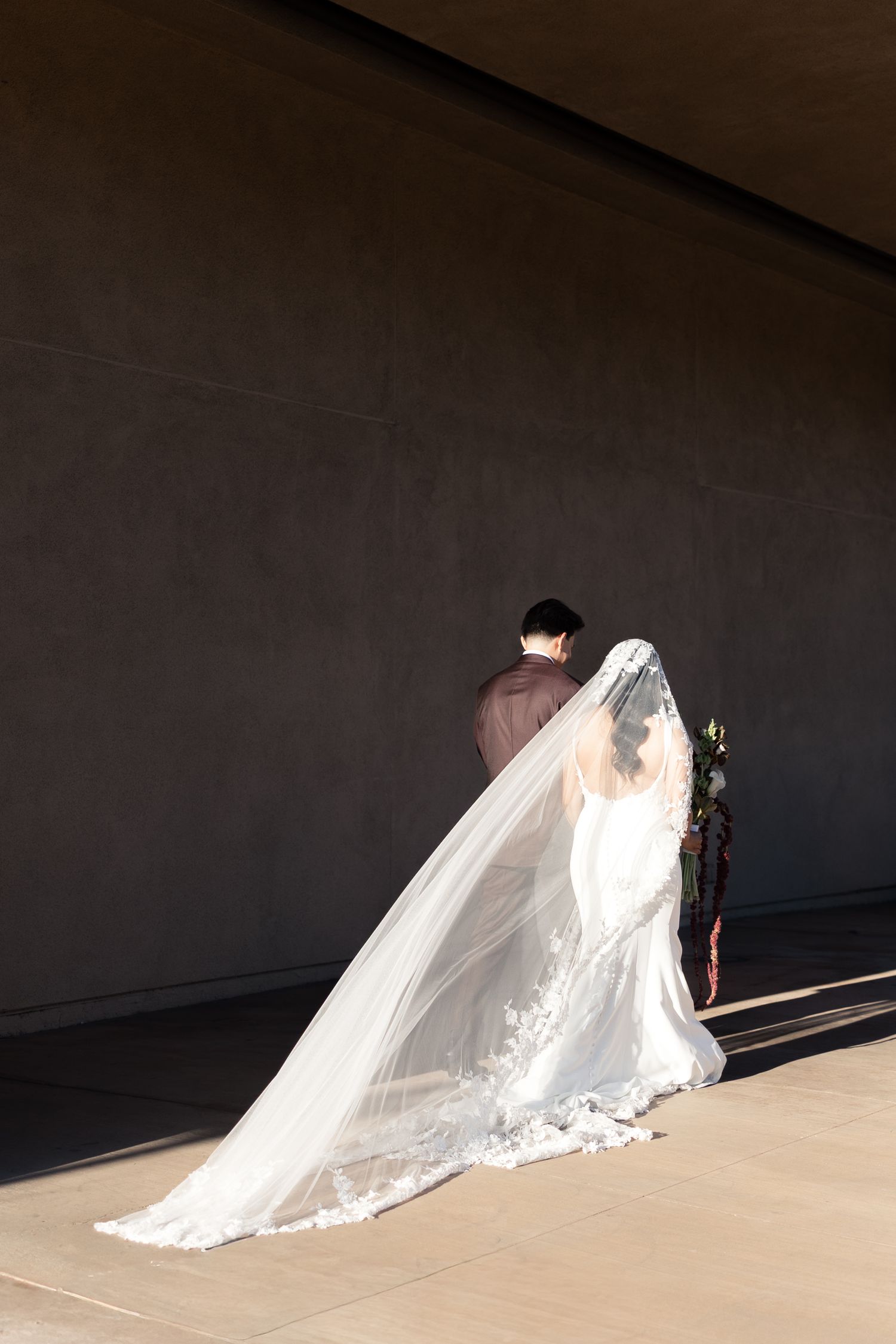 Bride in white lace gown with long cathedral veil stands with groom against gray wall in soft sunlight.