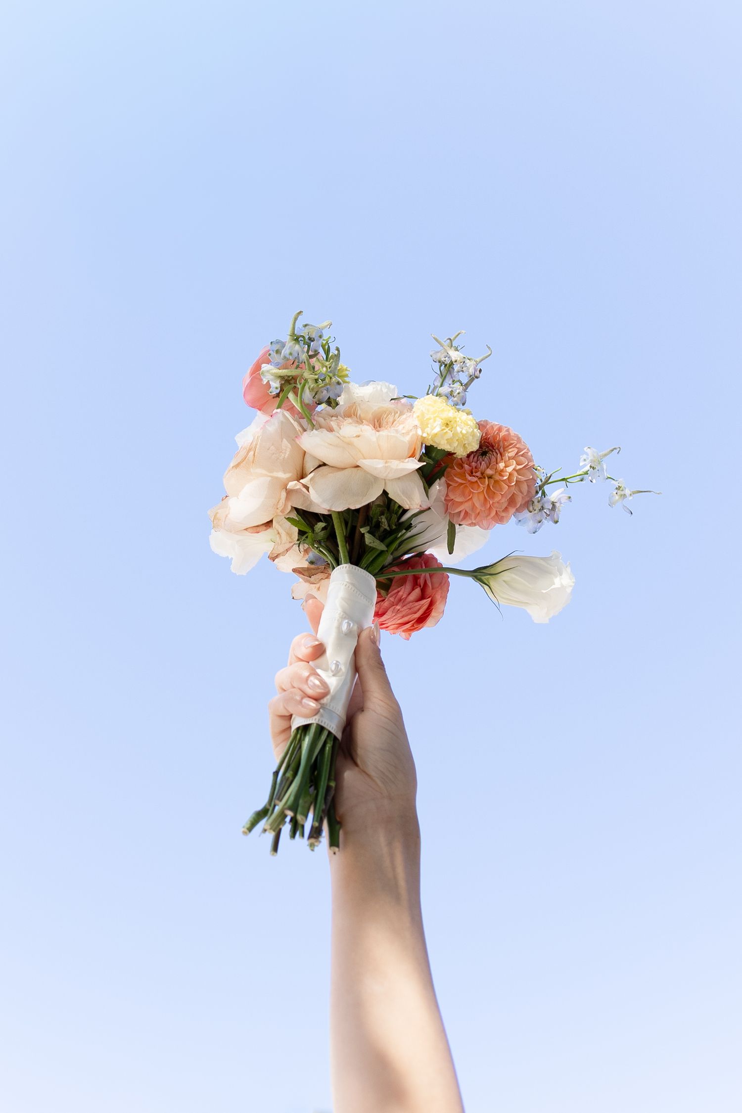 A hand raised against a blue sky holding a beautiful bouquet of white, pink, and coral flowers.