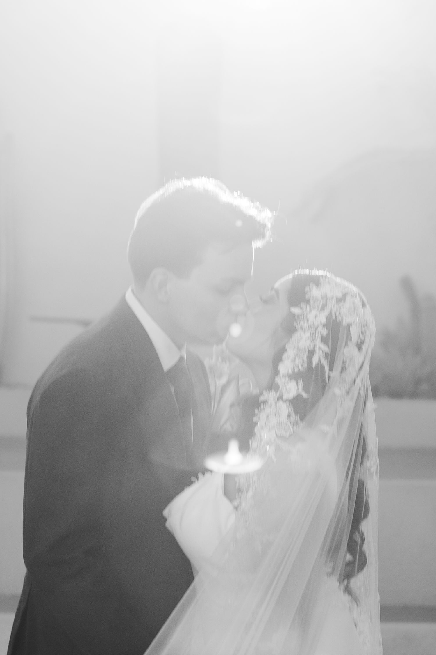 Black and white photo of a bride and groom sharing their first kiss at the altar, bride wearing lace veil.