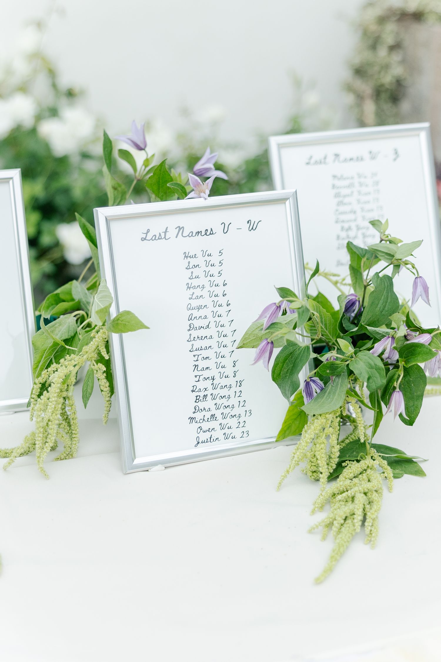 Elegant wedding seating chart in silver frame decorated with green ivy and purple flowers on a white table.