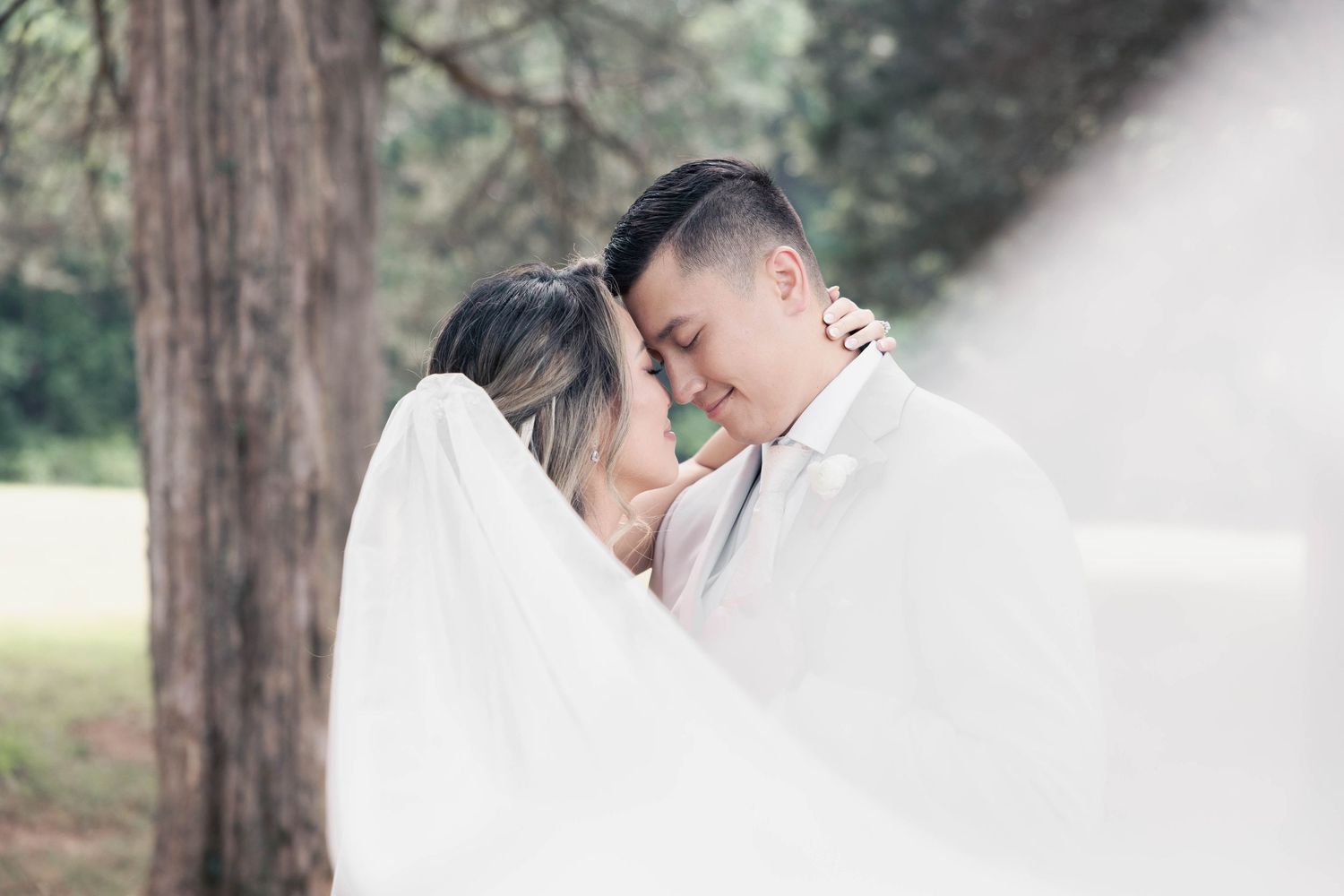 Romantic couple in white outfits embracing among tall trees in soft lighting.
