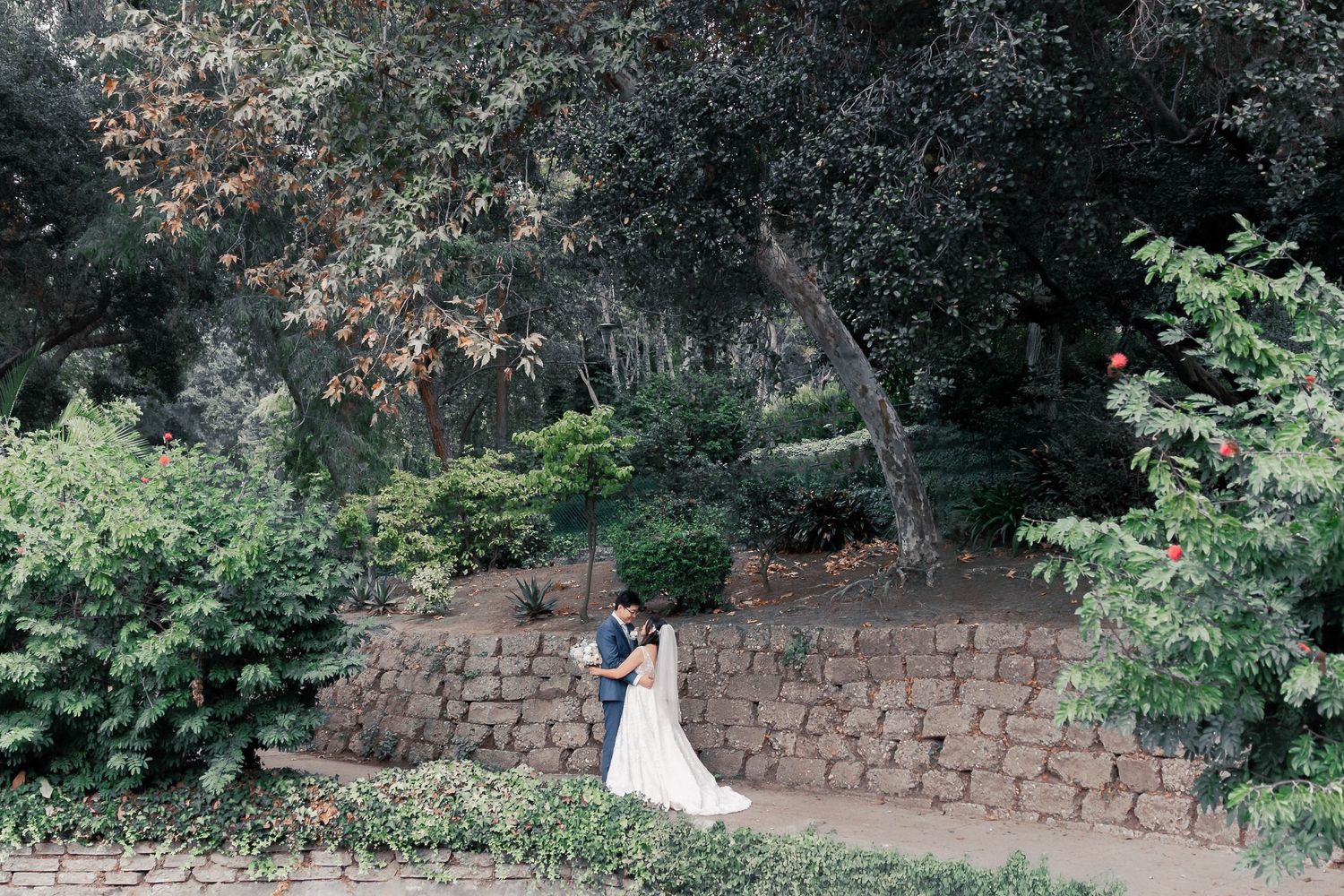 Couple in wedding attire poses against rustic stone wall in garden setting at dusk.