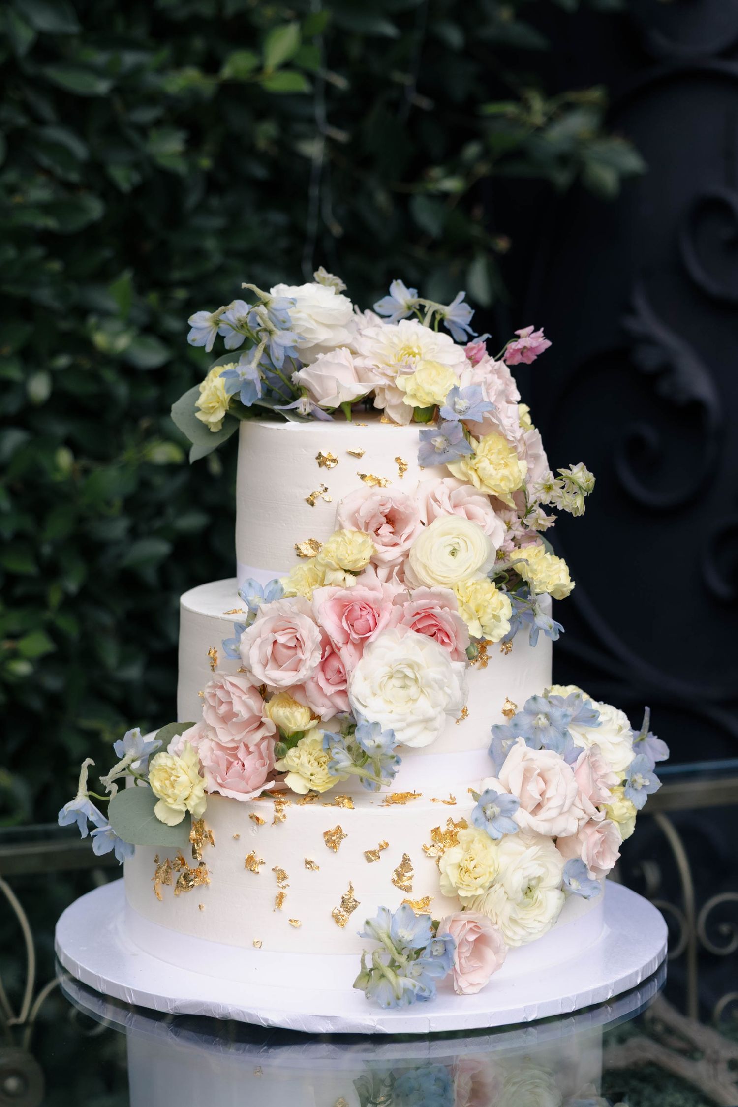 Elegant white wedding cake decorated with cascading pink roses, blue hydrangeas and delicate floral accents on display outdoors.