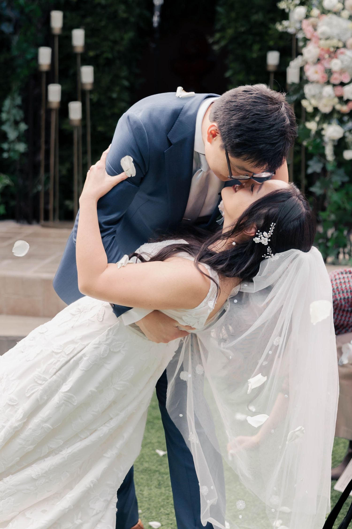 A romantic wedding couple shares a dip kiss in a garden setting with floral decorations and lanterns.