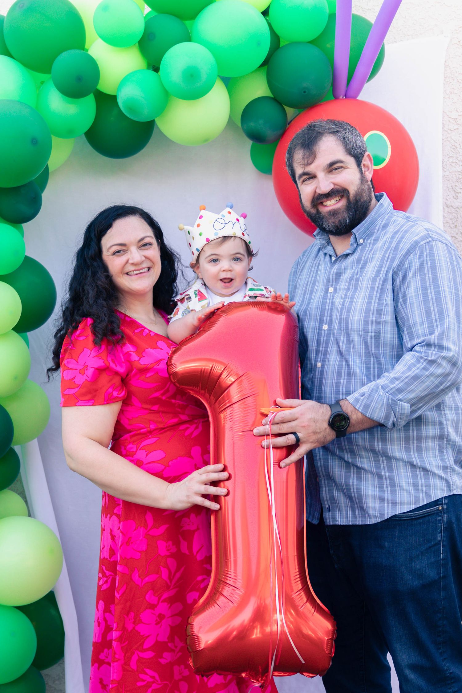 A family poses together against a green balloon arch backdrop celebrating a first birthday party with a red number one balloon.