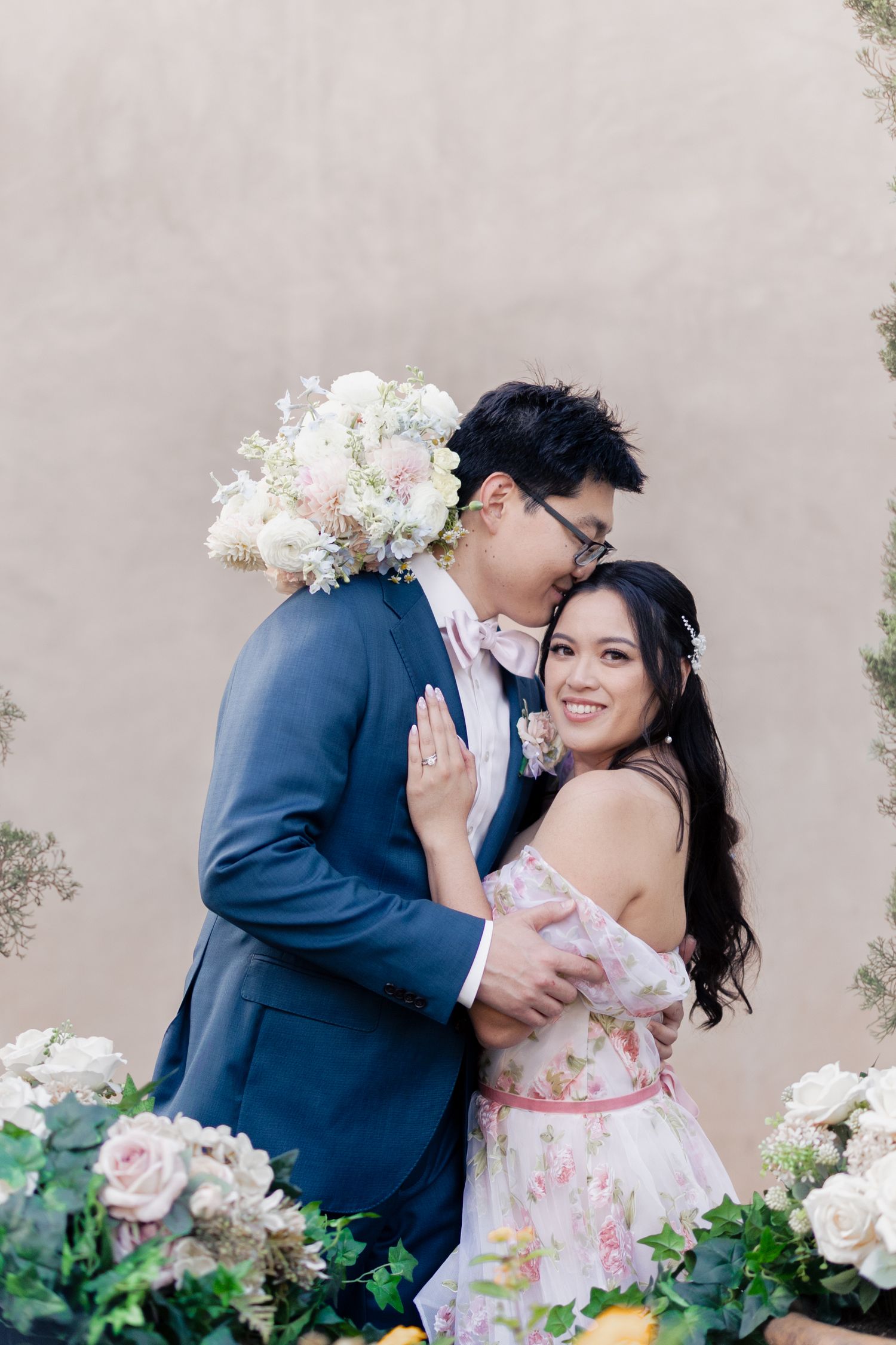 A romantic couple embraces in front of a beige wall with floral decorations, wearing formal attire for an engagement photo.