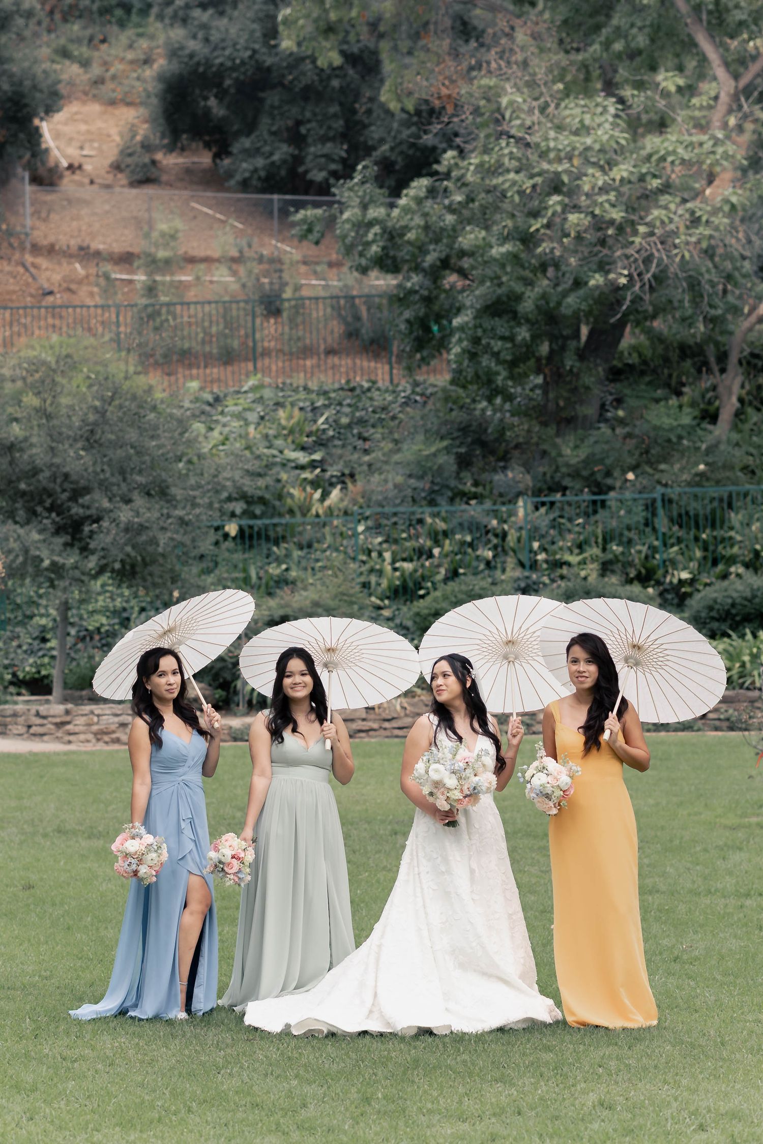 Wedding party in pastel dresses holding white parasols pose together on a grassy lawn with trees in background.