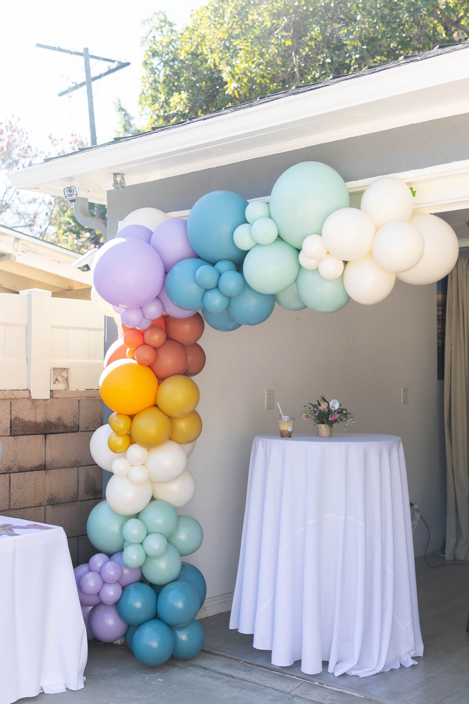 Elegant party setup with cascading balloon arch in pastel colors of blue, purple, and gold over white tablecloths.