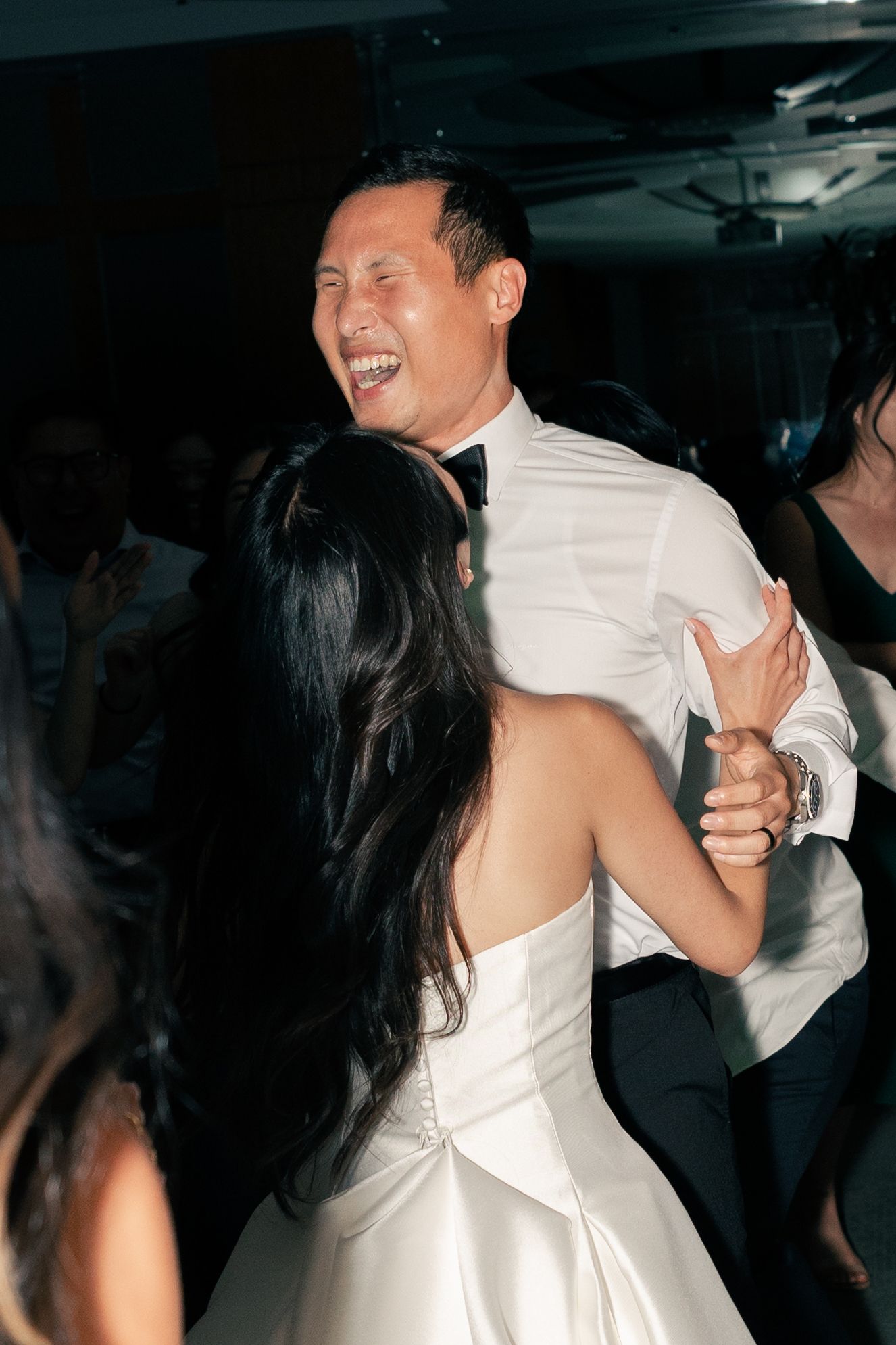 Bride and groom share a joyful first dance at their wedding reception, groom smiling broadly in white shirt and bow tie.