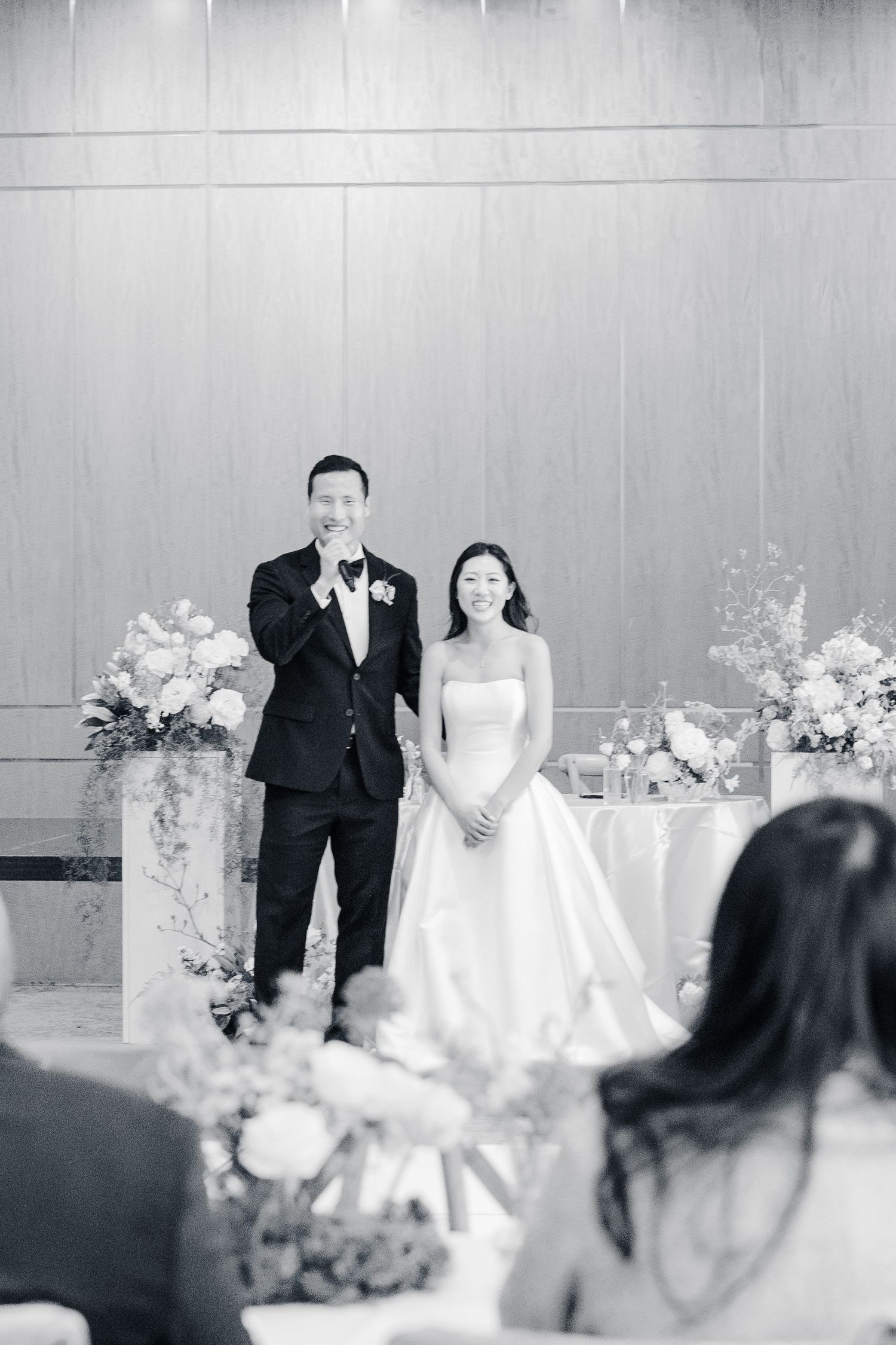 Black and white photo of a groom wiping tears and smiling bride standing at wedding altar with floral arrangements.