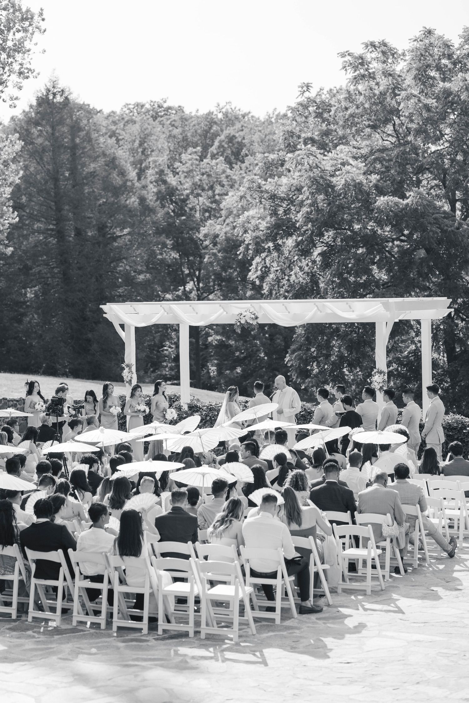 Black and white photo of outdoor wedding ceremony under white pergola with seated guests.