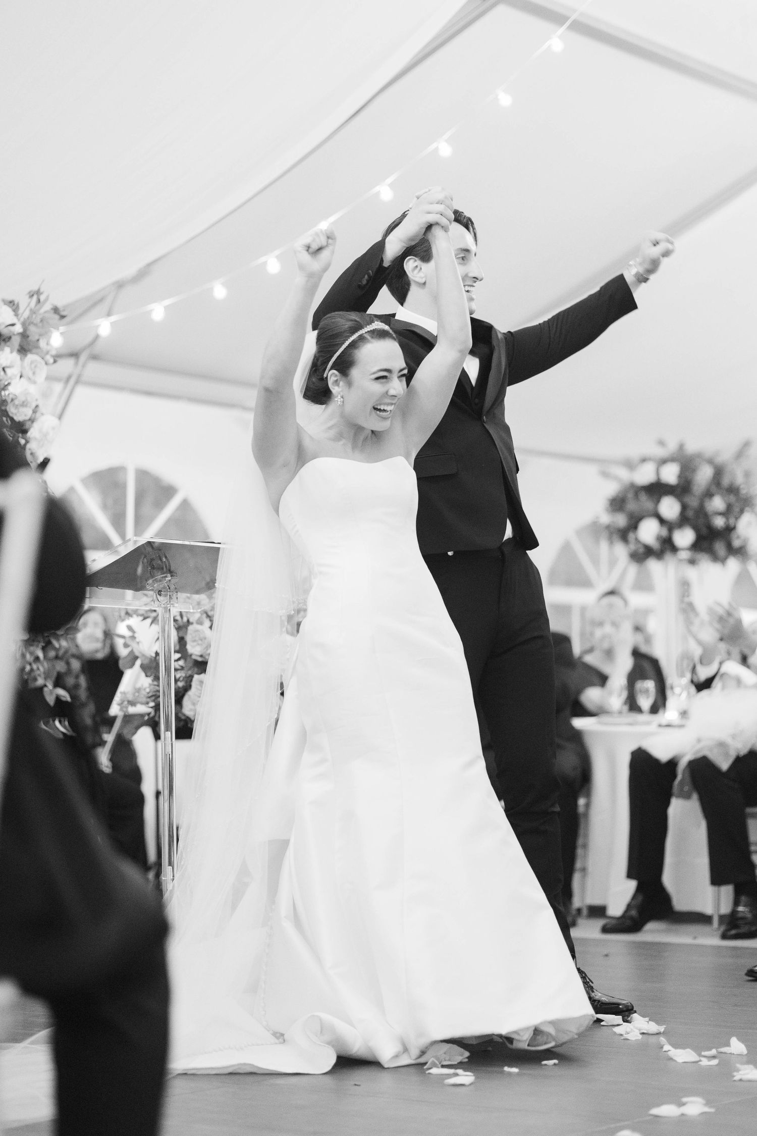 Black and white photo of a bride and groom sharing their first dance under a tent with string lights at their wedding reception.