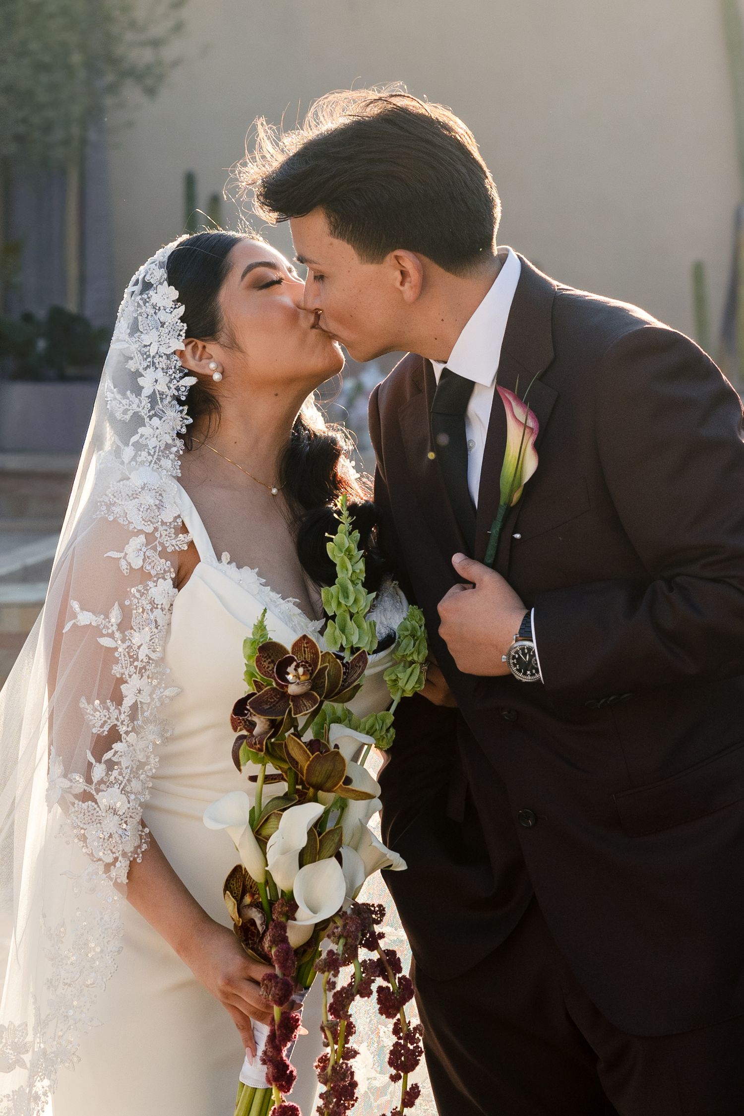 Bride and groom share a kiss, she wears a lace veil and holds a bouquet of dark florals and calla lilies.