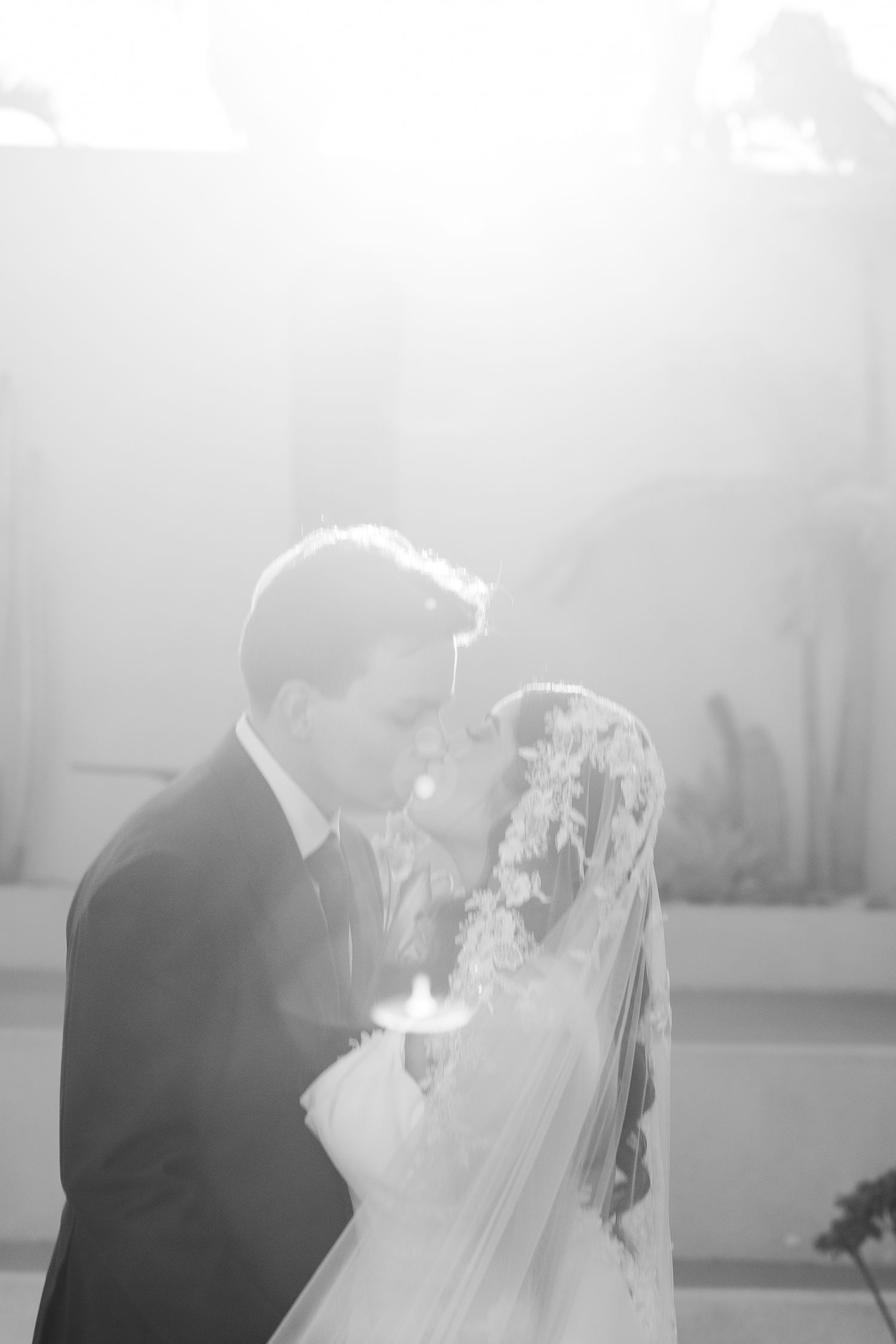 Black and white photo of a bride and groom sharing a romantic kiss, bathed in soft glowing light on their wedding day.