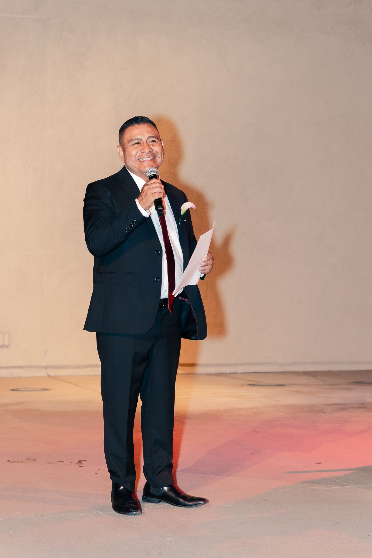 Man in black suit speaking into microphone while holding papers at a formal event.