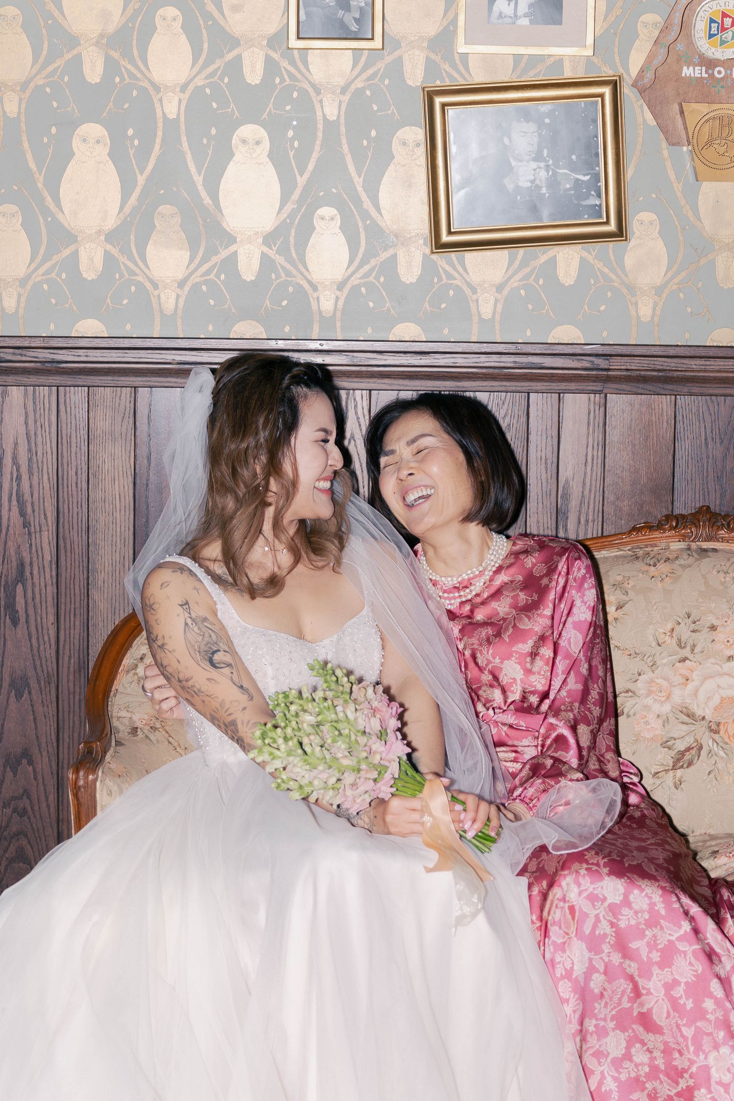 Two people sharing joyful laughter on an antique couch, one in a wedding dress holding a bouquet.