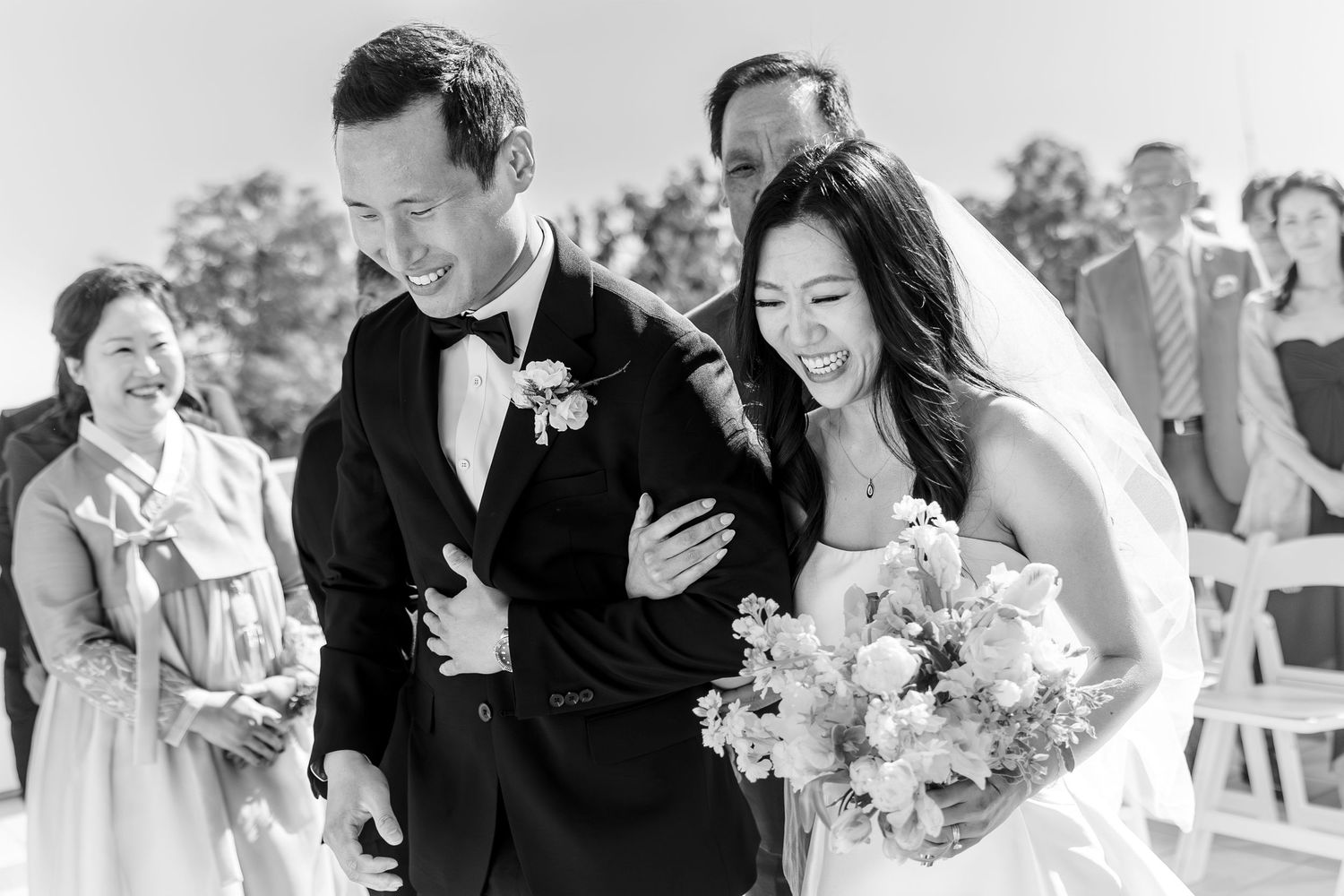 Happy newlyweds laugh joyfully after outdoor wedding ceremony, bride holding bouquet, groom in tuxedo, guests smiling behind them.