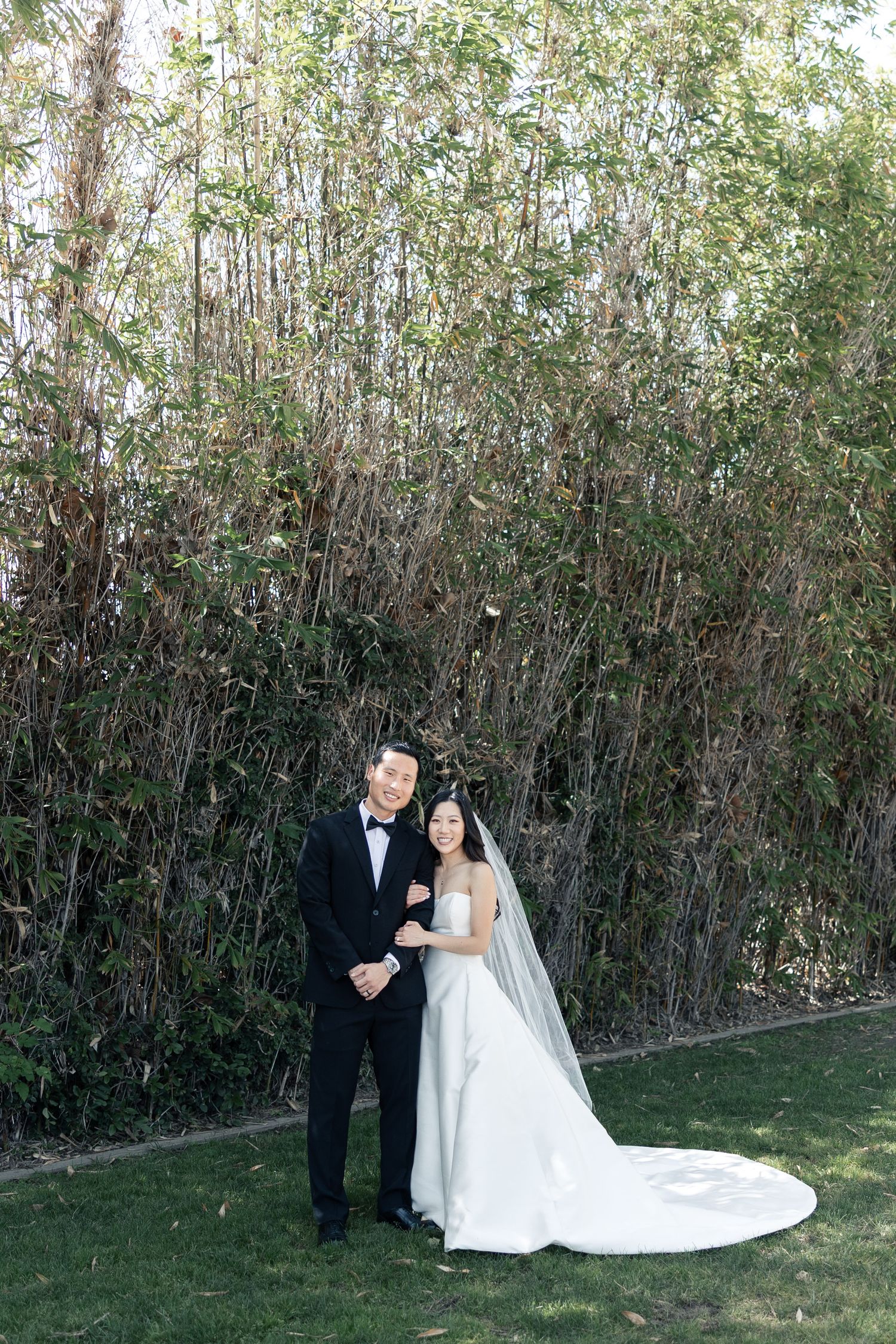 Bride and groom pose together in front of lush bamboo greenery, bride wearing a white gown with long veil.