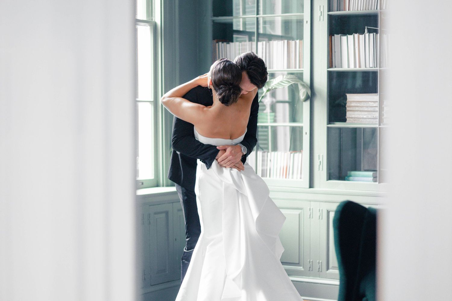 Bride and groom sharing romantic embrace in elegant library, bride in stunning white backless wedding gown.
