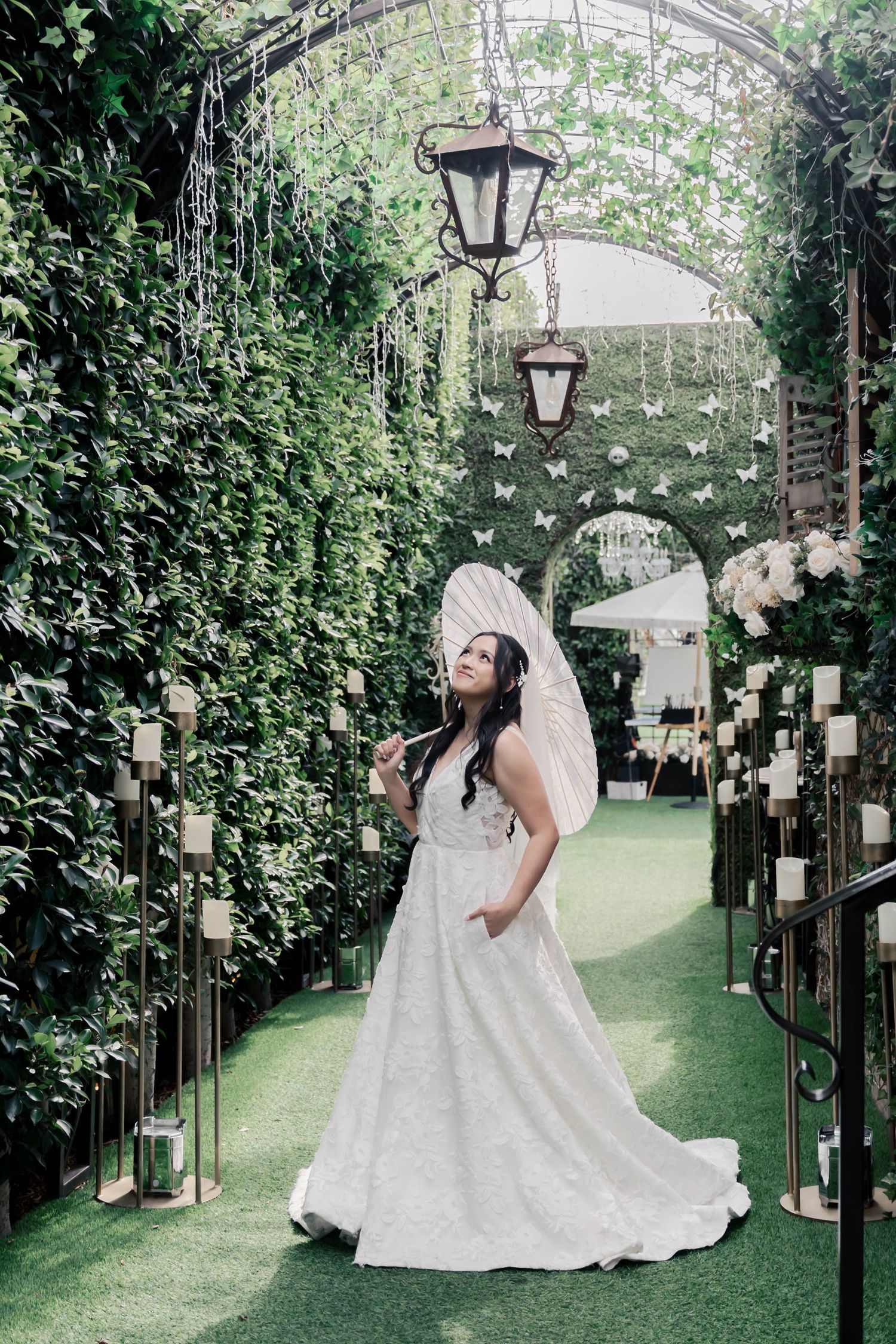 A bride in a flowing white wedding gown poses gracefully in a garden setting with vintage lamp posts and greenery.