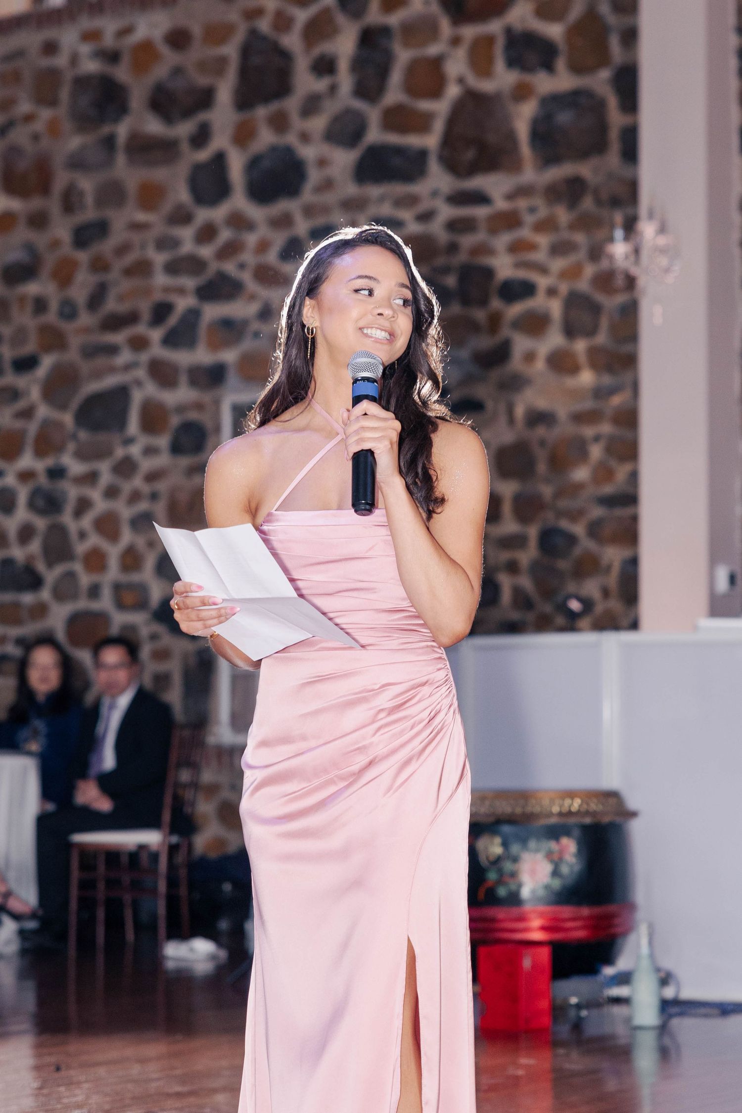 A member of the wedding party in pink dress gives a speech at stone-walled reception venue.