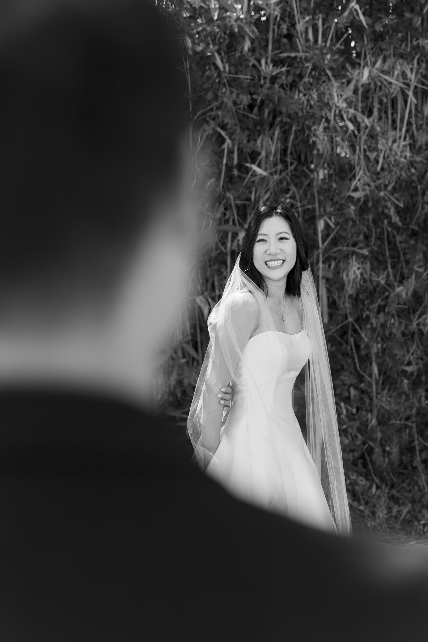 Black and white photo of smiling Asian bride in strapless gown and veil during outdoor first look moment.