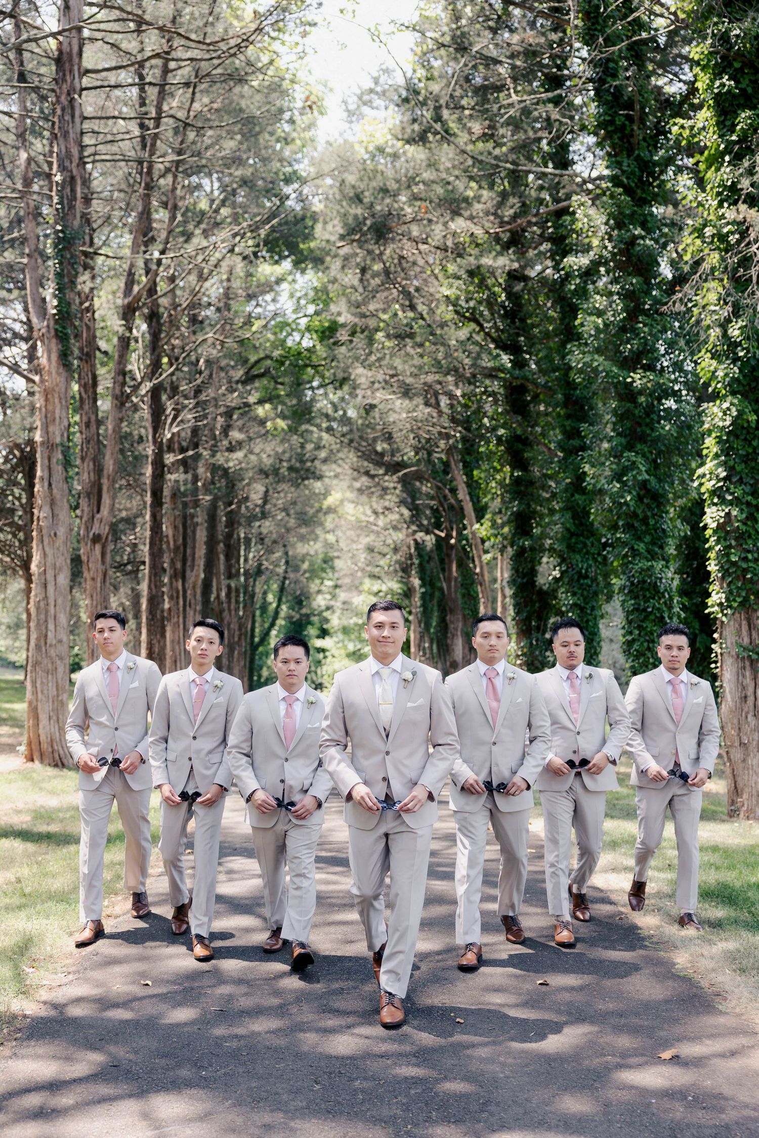 Wedding party members in matching light gray suits pose playfully on a wooded path.